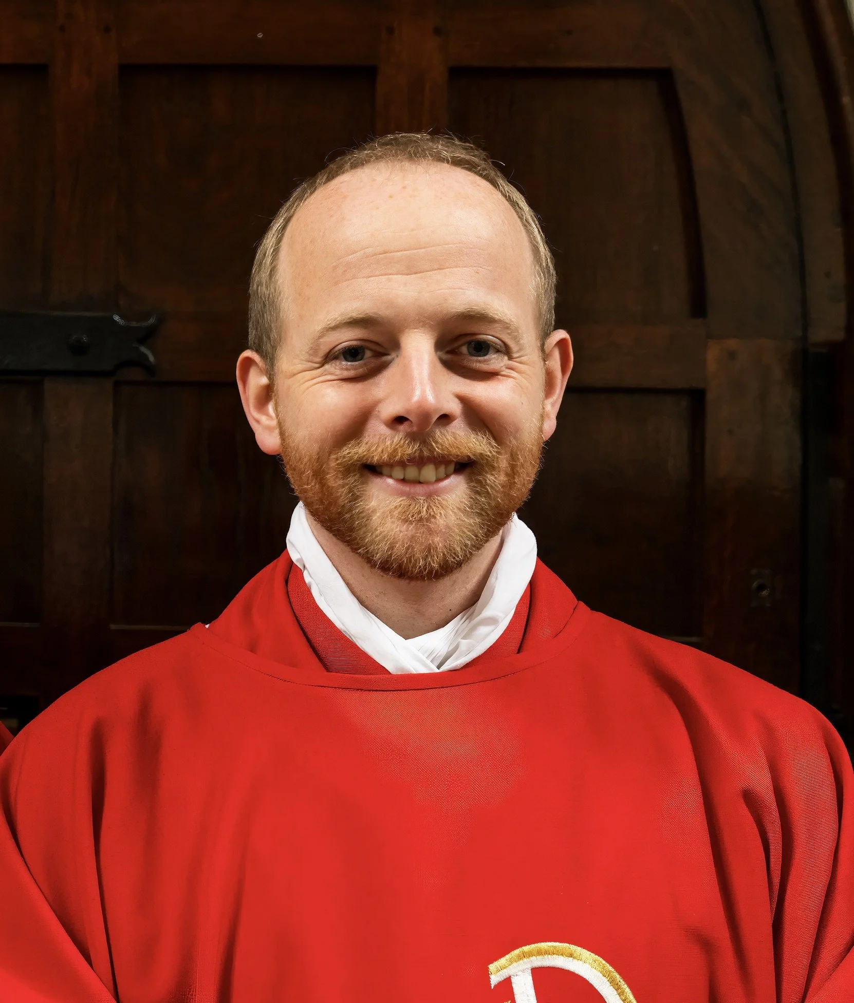 A smiling young man in a black clerical shirt with a white collar, wearing a black suit jacket, standing indoors with a blurred wooden interior background.