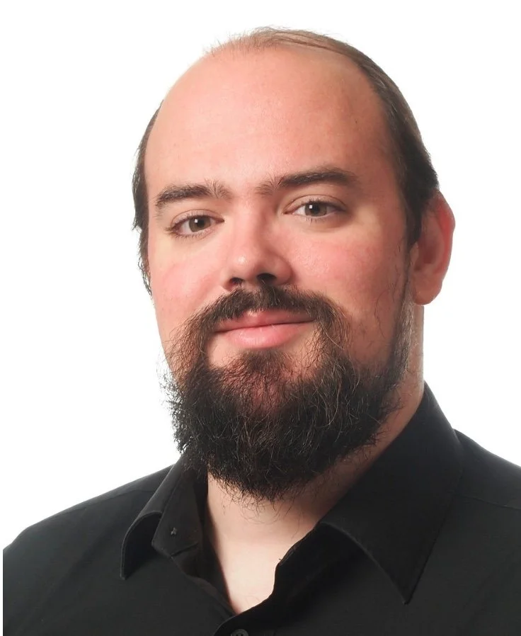 A man with a beard and receding hairline wearing a black collared shirt, smiling slightly, against a plain white background.