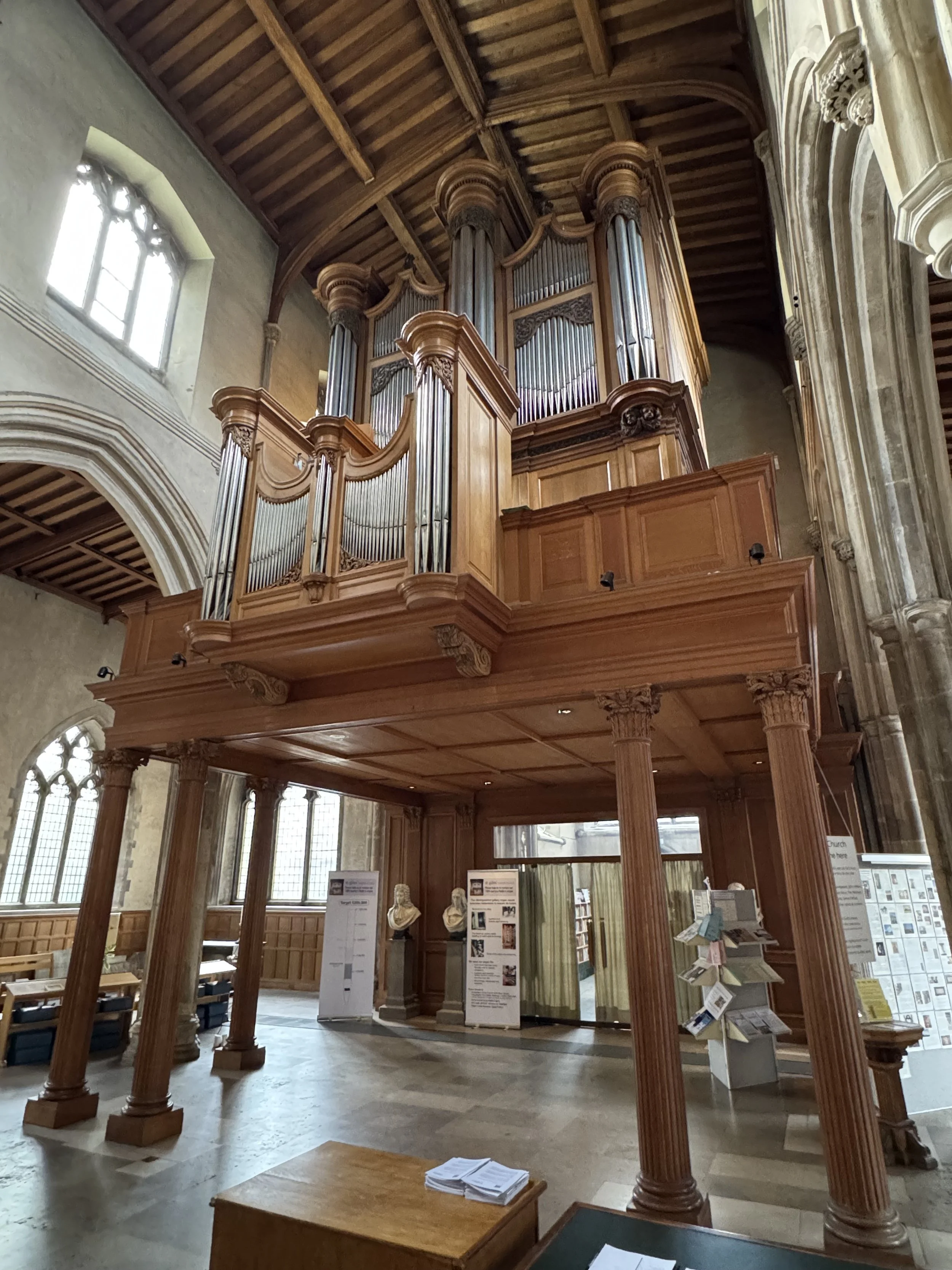 Interior of a church with a large wooden pipe organ on a raised platform, flanked by classical columns, high arched windows, and informational signs and bust sculptures in the foreground.