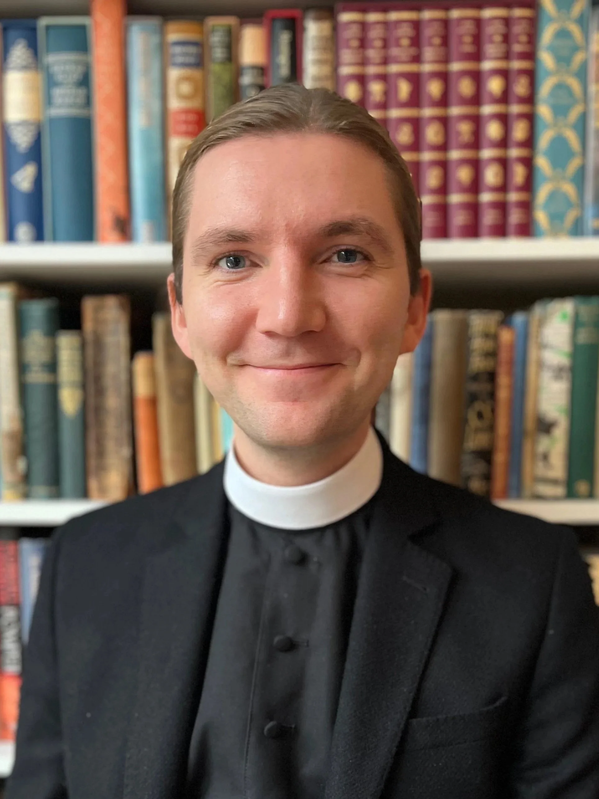 Smiling young man with short light brown hair, wearing a clerical collar and a gray suit jacket, outdoors with blurred green foliage in the background.