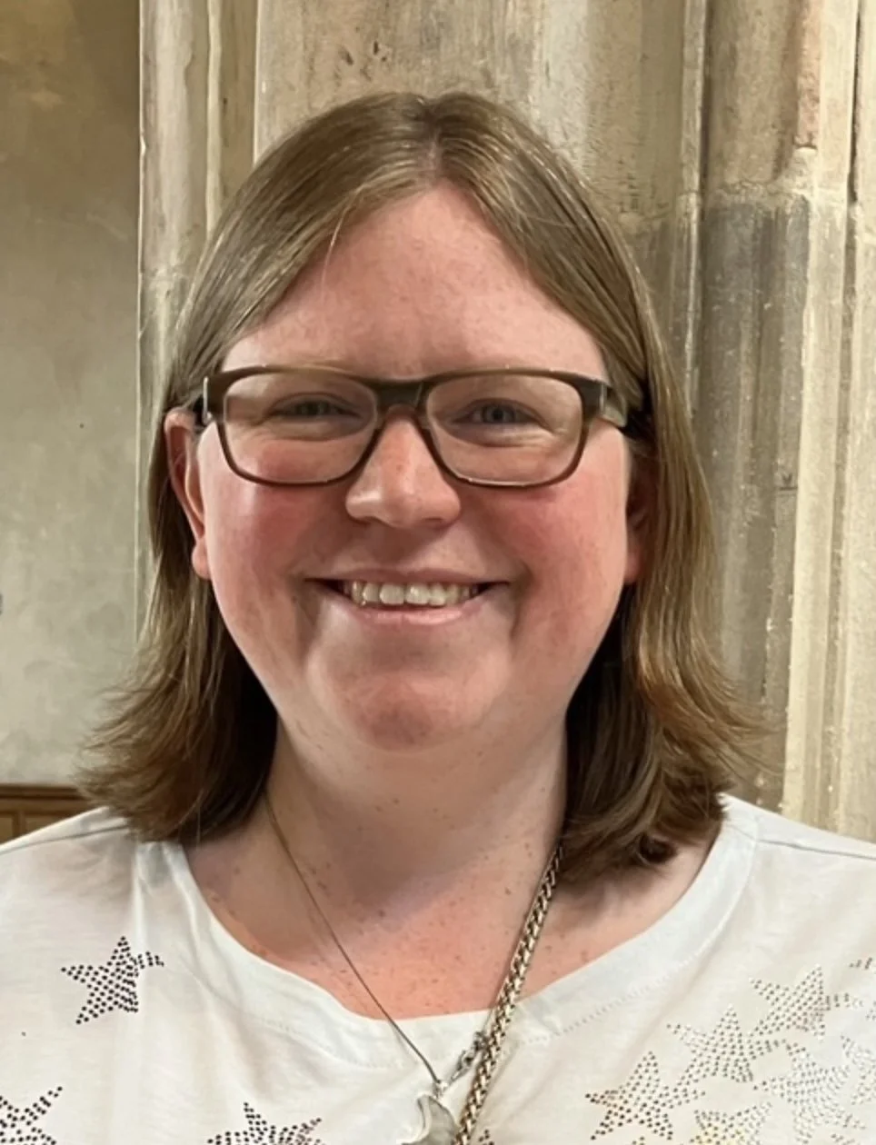 Smiling woman with glasses, shoulder-length brown hair, wearing a white shirt with star patterns, standing indoors with stone columns in the background.