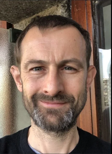 Close-up of a smiling man with short dark hair, a beard with gray, and blue eyes, standing outdoors near a building with wood and glass elements.