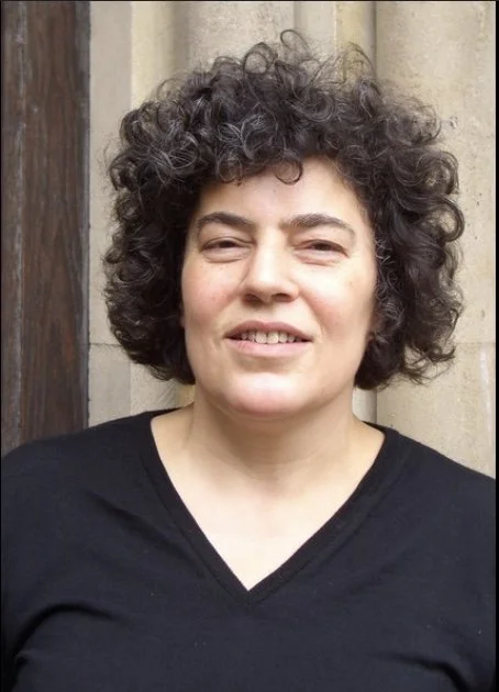 A woman with curly dark hair and light skin, wearing a black V-neck shirt, standing outdoors in front of a stone wall.