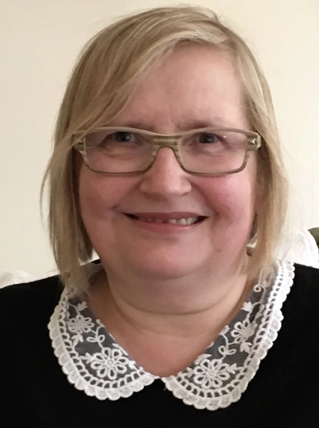 A woman with blonde hair, glasses, and a black top with a white embroidered collar, smiling in front of a plain light-colored background.