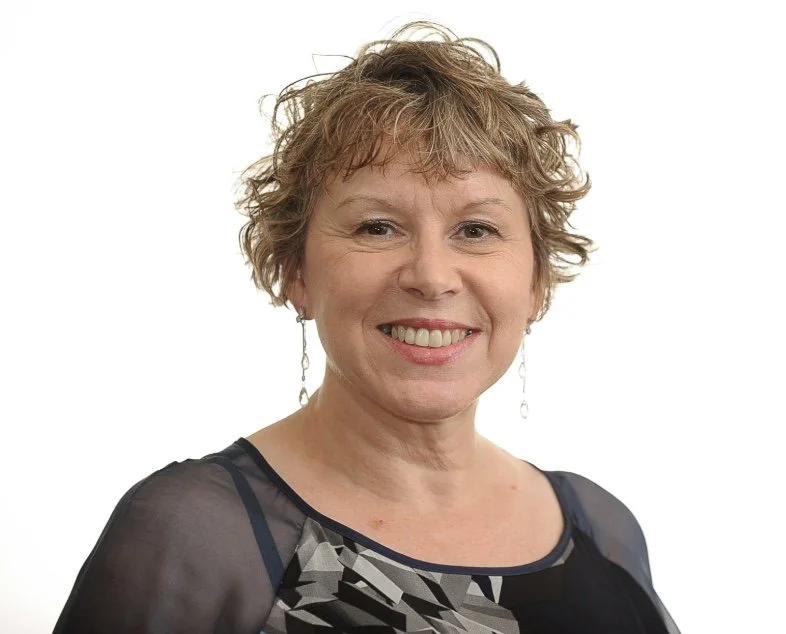 A middle-aged woman with short, curly blonde hair, wearing a black and gray patterned top and drop earrings, smiling against a plain white background.