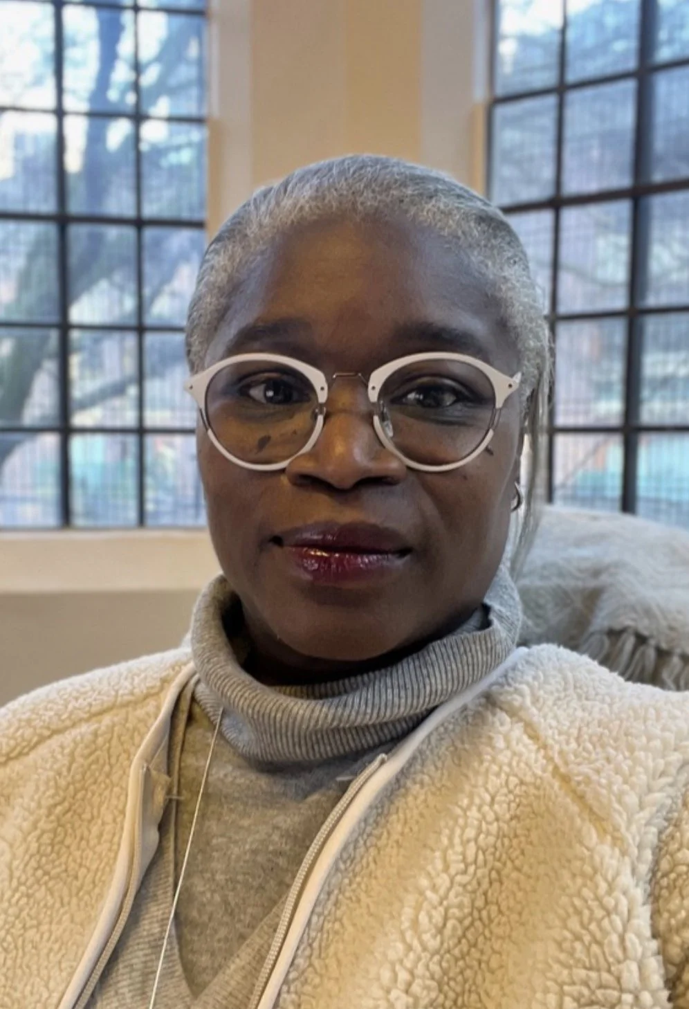 A woman with short gray hair and glasses sitting indoors near large windows.