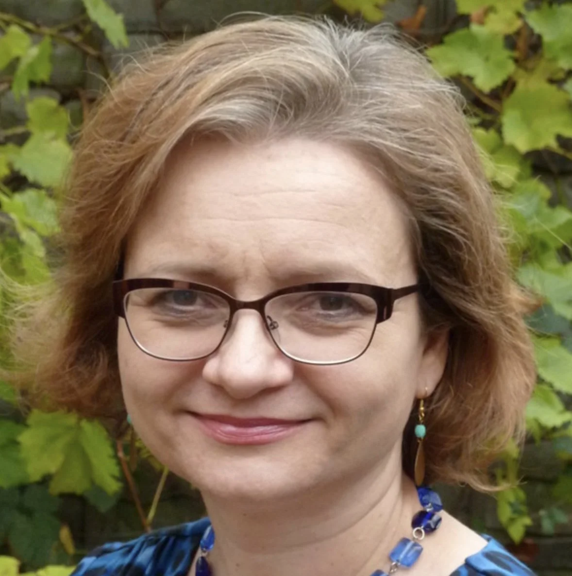Close-up of a woman with short curly reddish-brown hair wearing glasses, blue jewelry, and a blue patterned top, outdoors in front of green foliage.