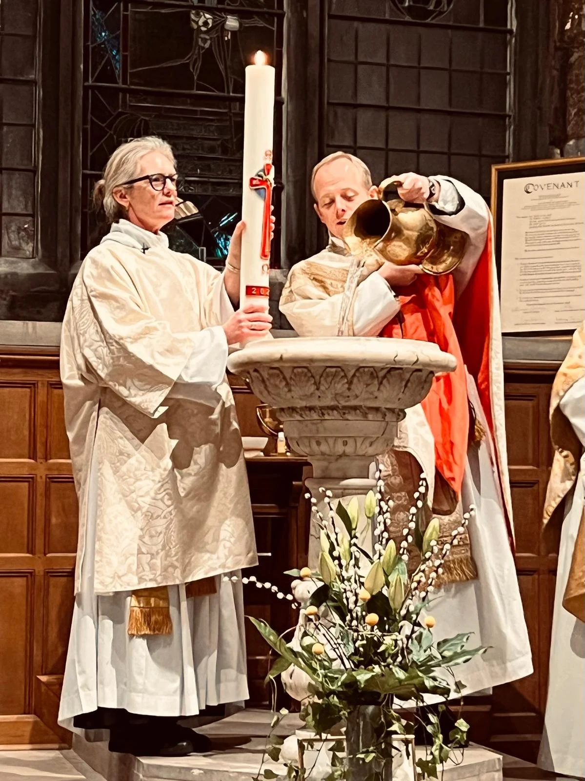 Clergy members performing a religious baptism ceremony inside a church, with a woman holding a large lit candle and a man pouring water from a pitcher into a baptismal font.