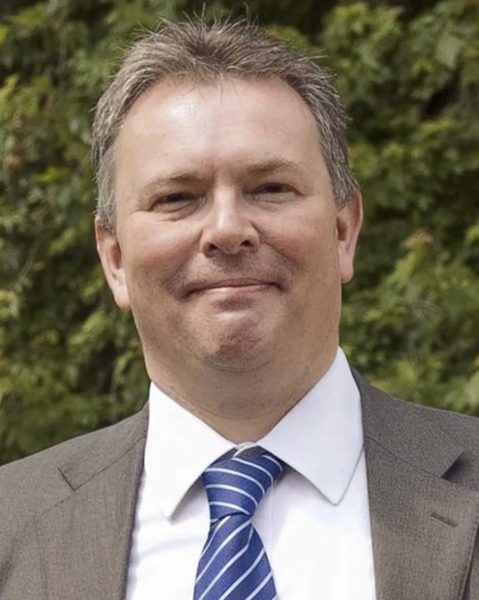 A man in a suit and tie standing outdoors with green trees in the background.