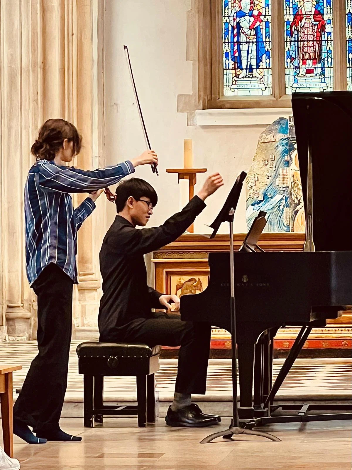 A young girl conducting an orchestra with a baton while a young man plays the piano in a church with stained glass windows.