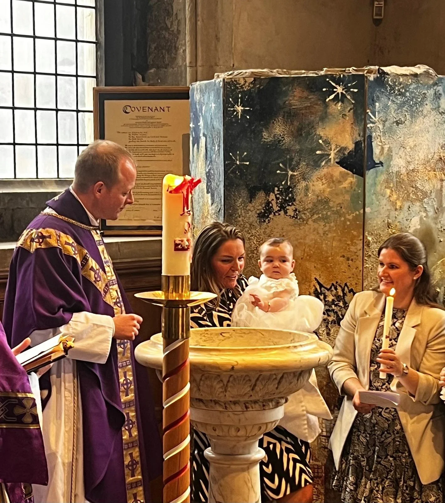 Baptism ceremony inside a church with a priest and a woman holding a baby in white clothes, another woman holding a lit candle, and a large decorated candle on a stand.