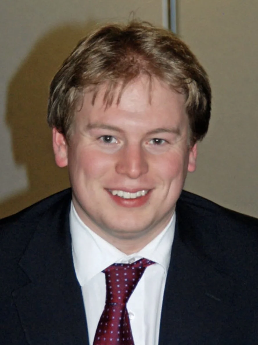 A young man with light brown hair, wearing a dark suit, white shirt, and a red tie, smiling at the camera indoors.