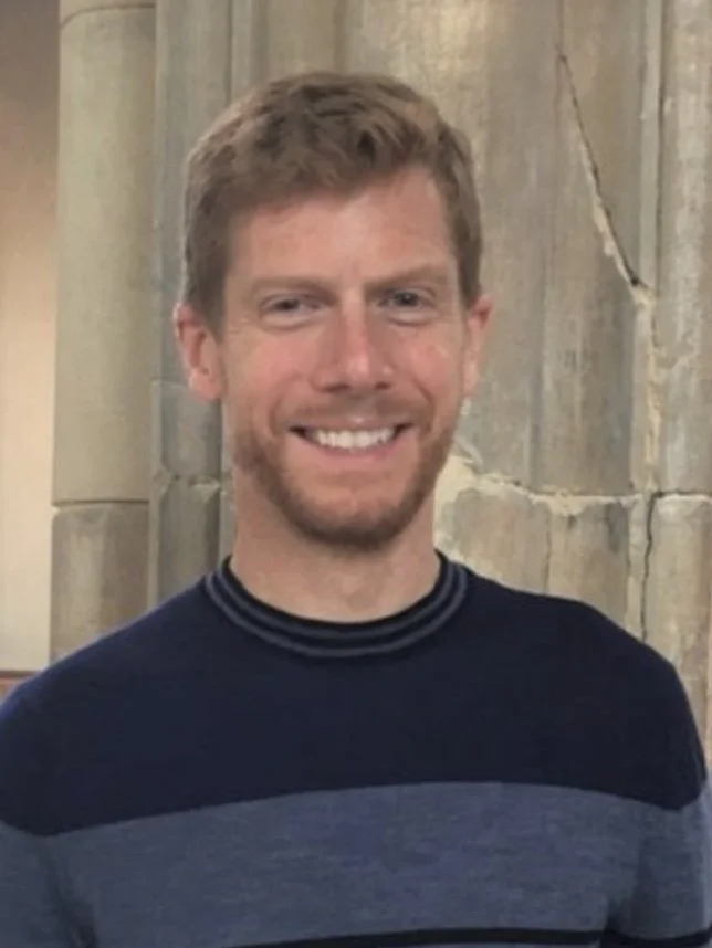A man with light brown hair, a beard, and a mustache is smiling at the camera. He is wearing a dark sweater with light and dark horizontal stripes and is standing in front of a stone wall.