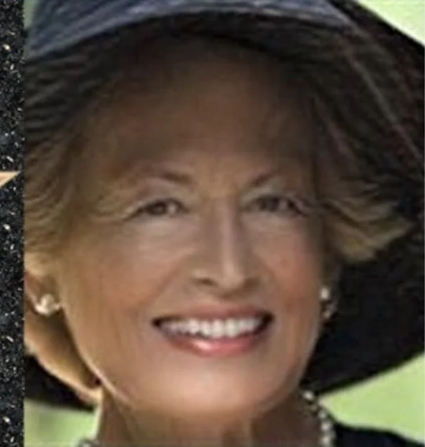 A woman smiling, wearing a large black hat and pearl earrings, outdoors in a sunny setting.