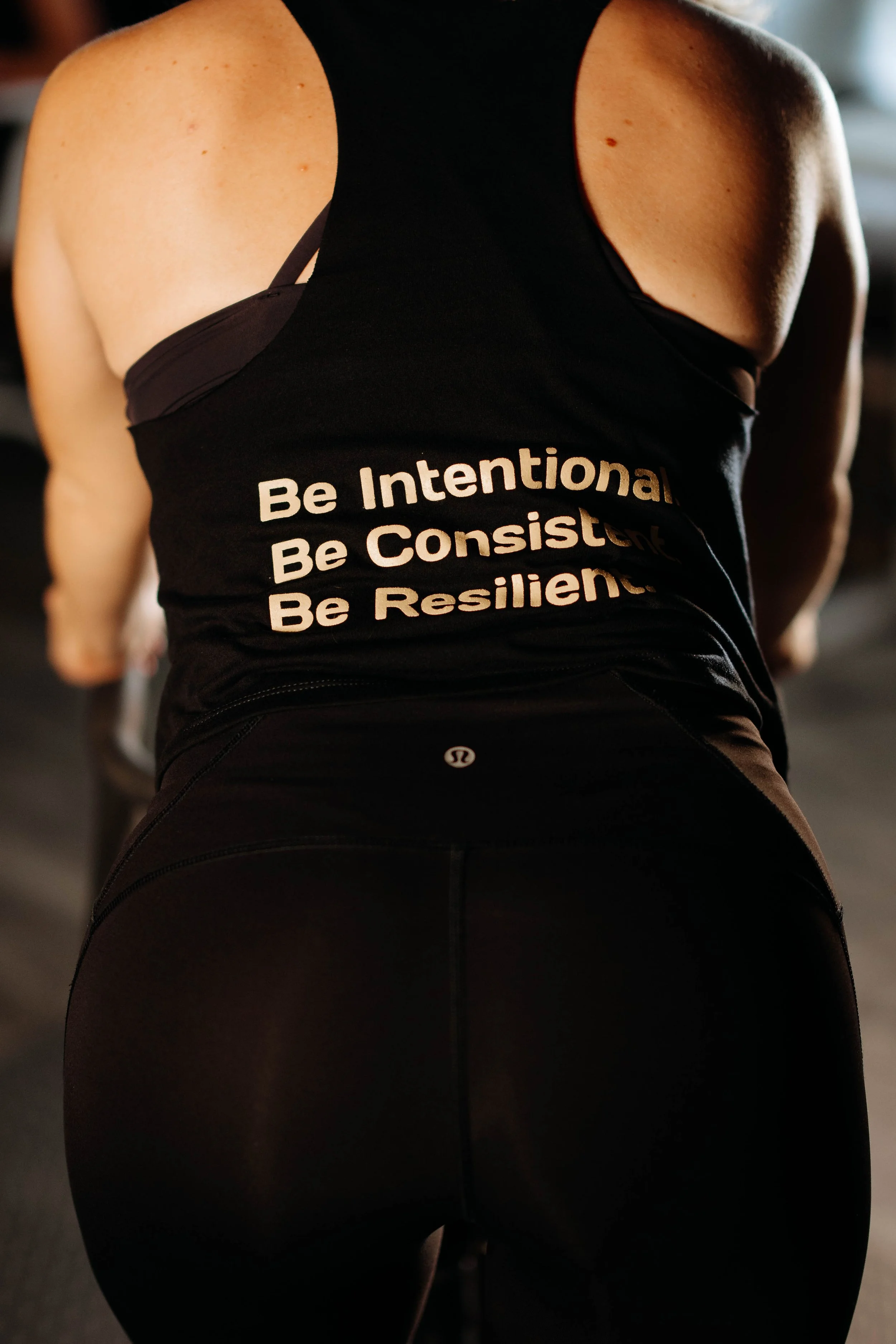 Close-up of a person wearing a black workout tank top with motivational text and black athletic leggings in a gym.
