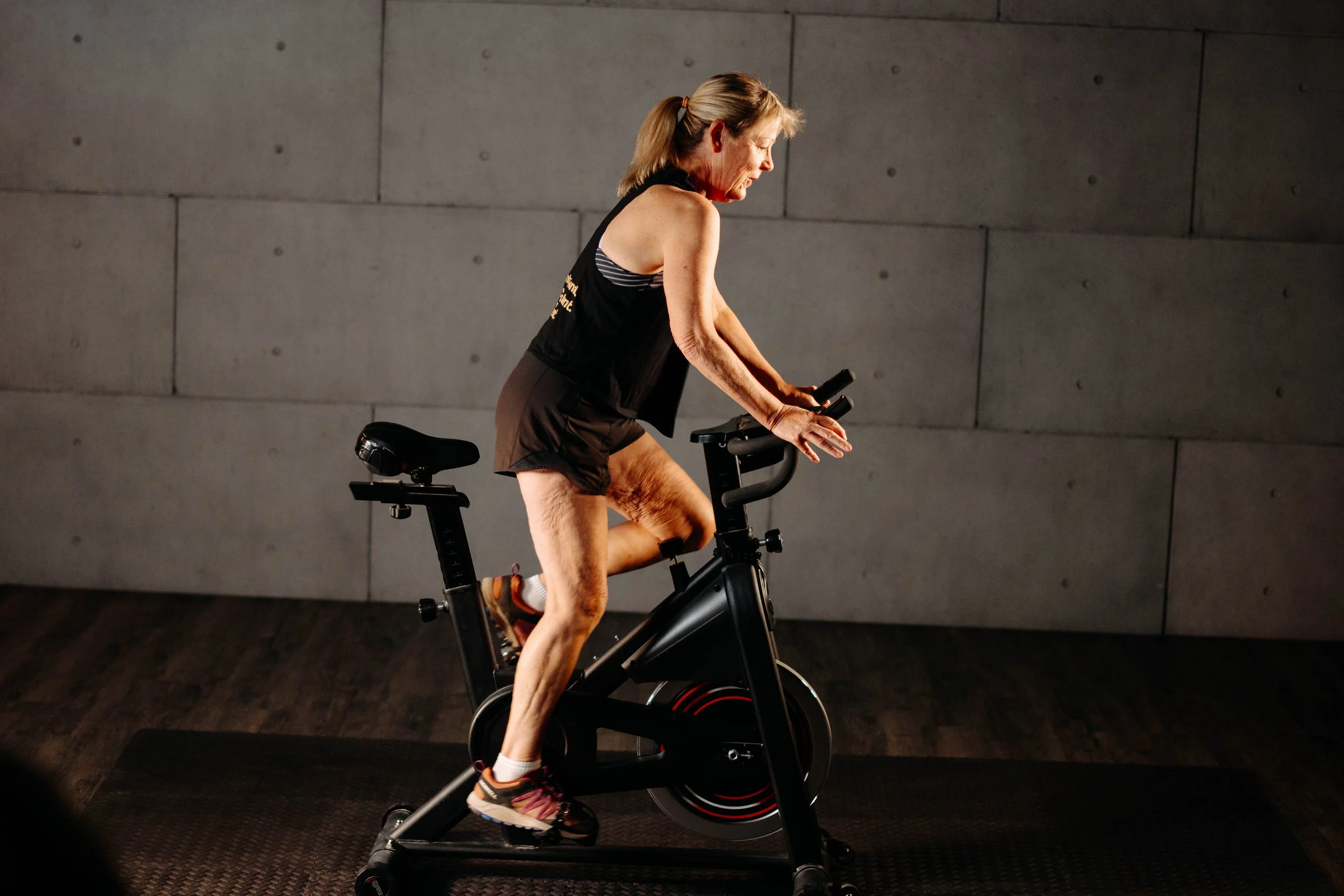 Older woman in workout clothes exercises on stationary bike indoors against a plain concrete wall.