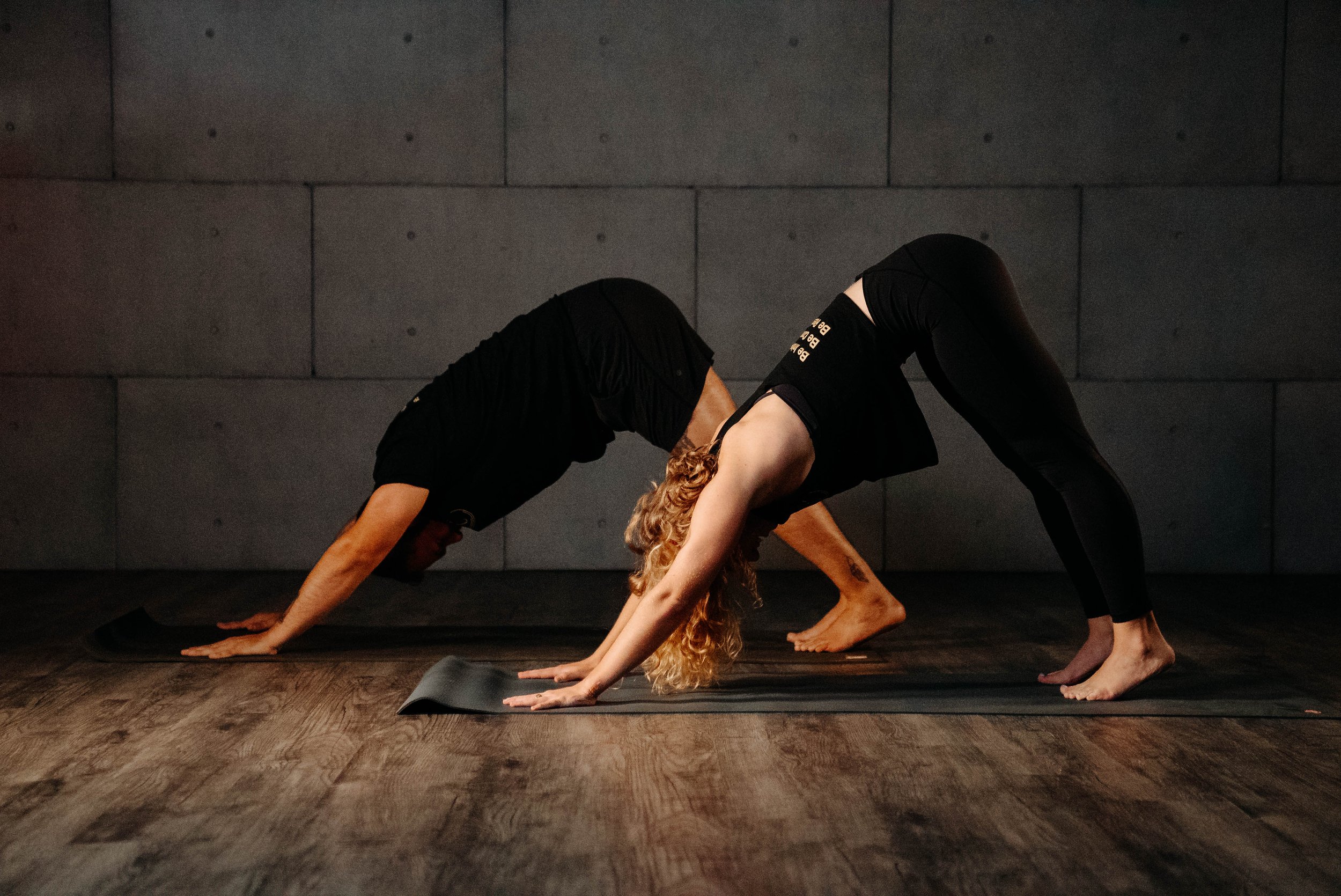 Two women practicing yoga in a dimly lit room with wooden floors and a concrete wall, both in downward dog pose.