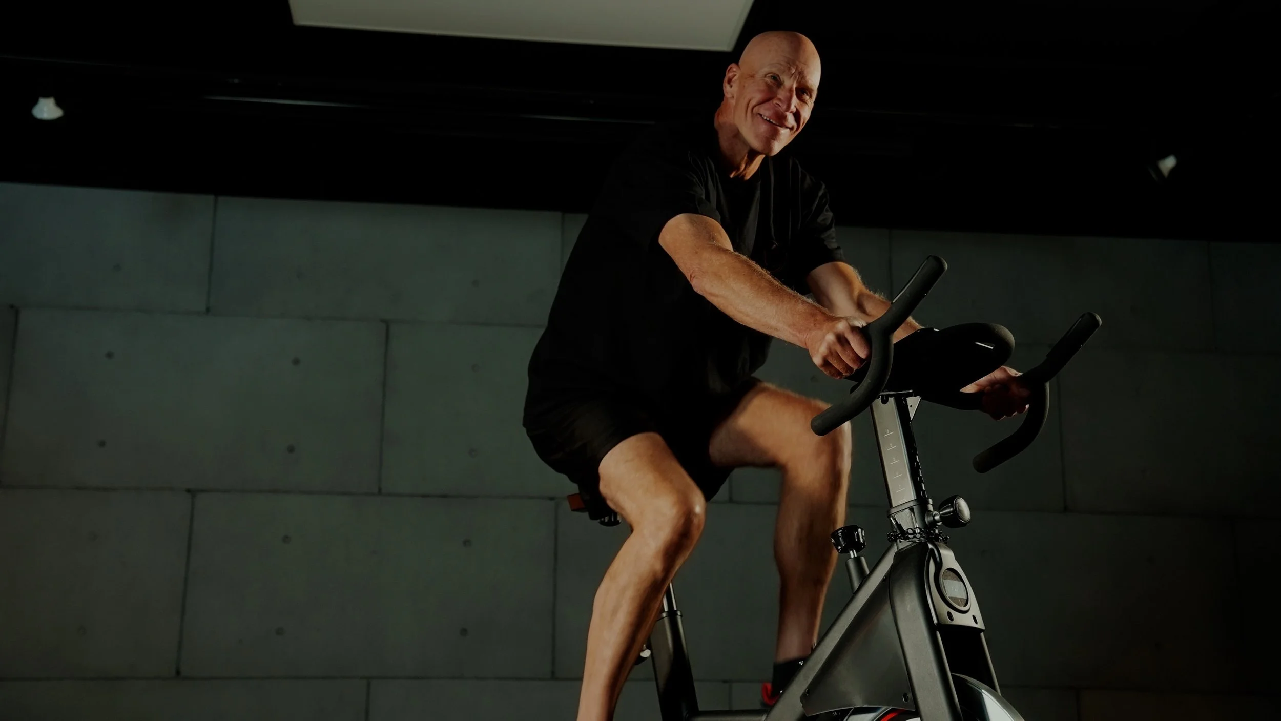 A man with a bald head and wearing a black shirt and shorts riding a stationary exercise bike indoors, smiling.