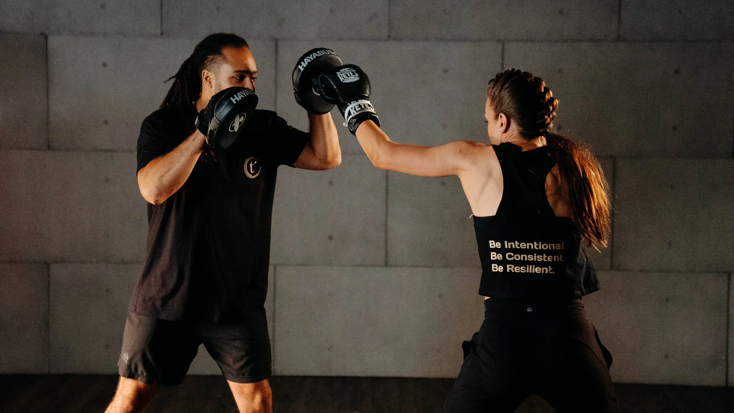 A woman wearing a black tank top with the words 'Be Intentional. Be Consistent. Be Resilient.' is boxing with a trainer or partner, who is wearing a black shirt and boxing gloves, in a gym with a dark wall in the background.
