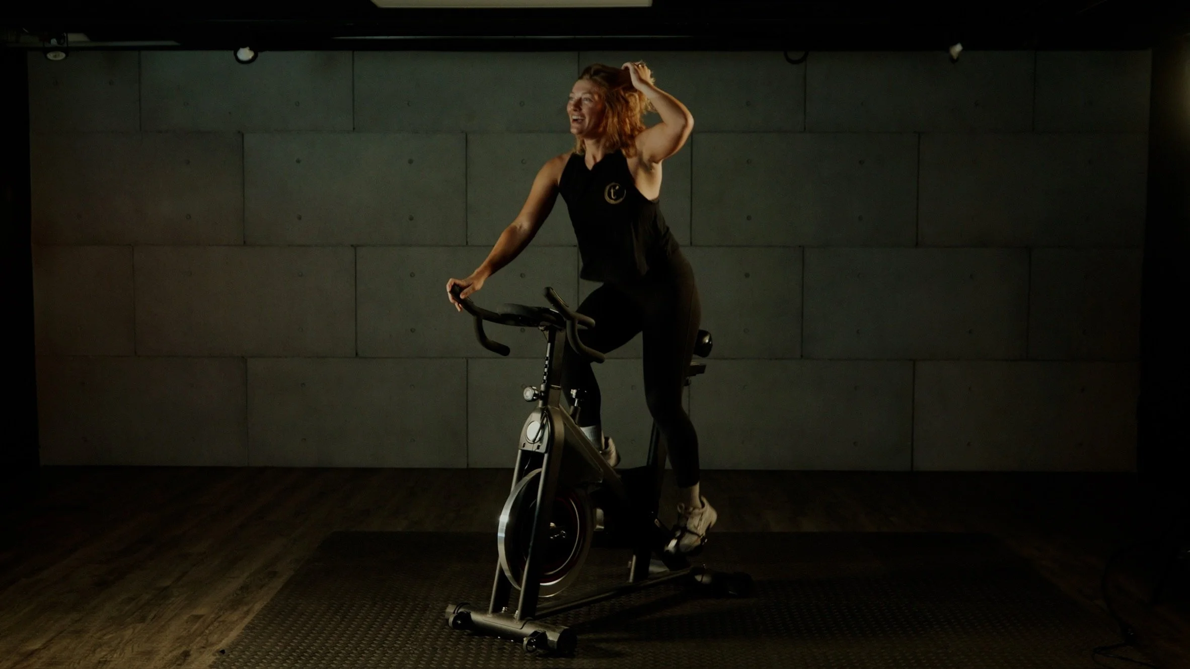 A woman with red hair riding a stationary exercise bike indoors, smiling and holding her head with one hand, with a dark plain wall behind her.