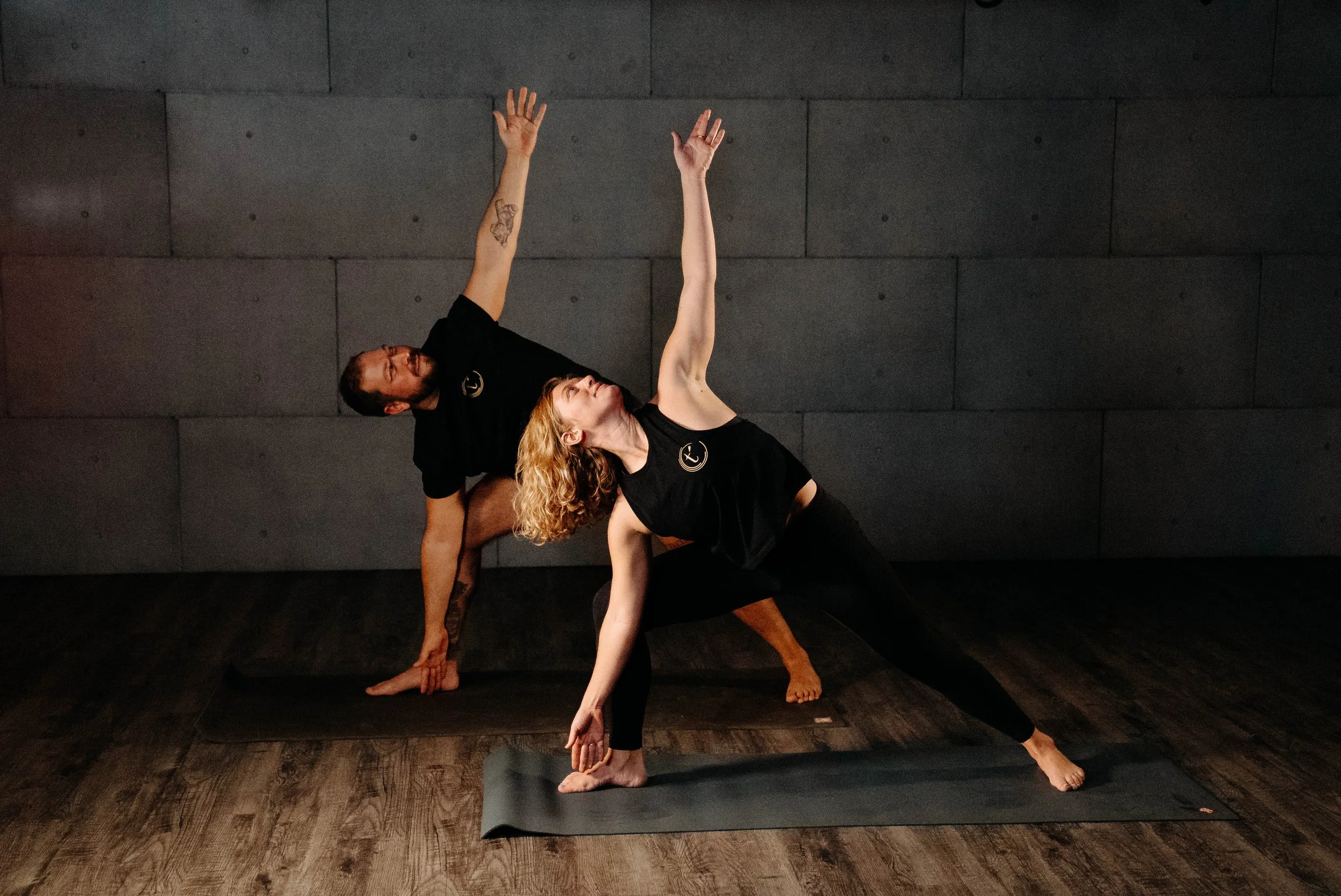 Two people practicing yoga on yoga mats in a dimly lit room. One person is in a side plank pose, while the other is in a modified side stretch pose, both facing the same direction.