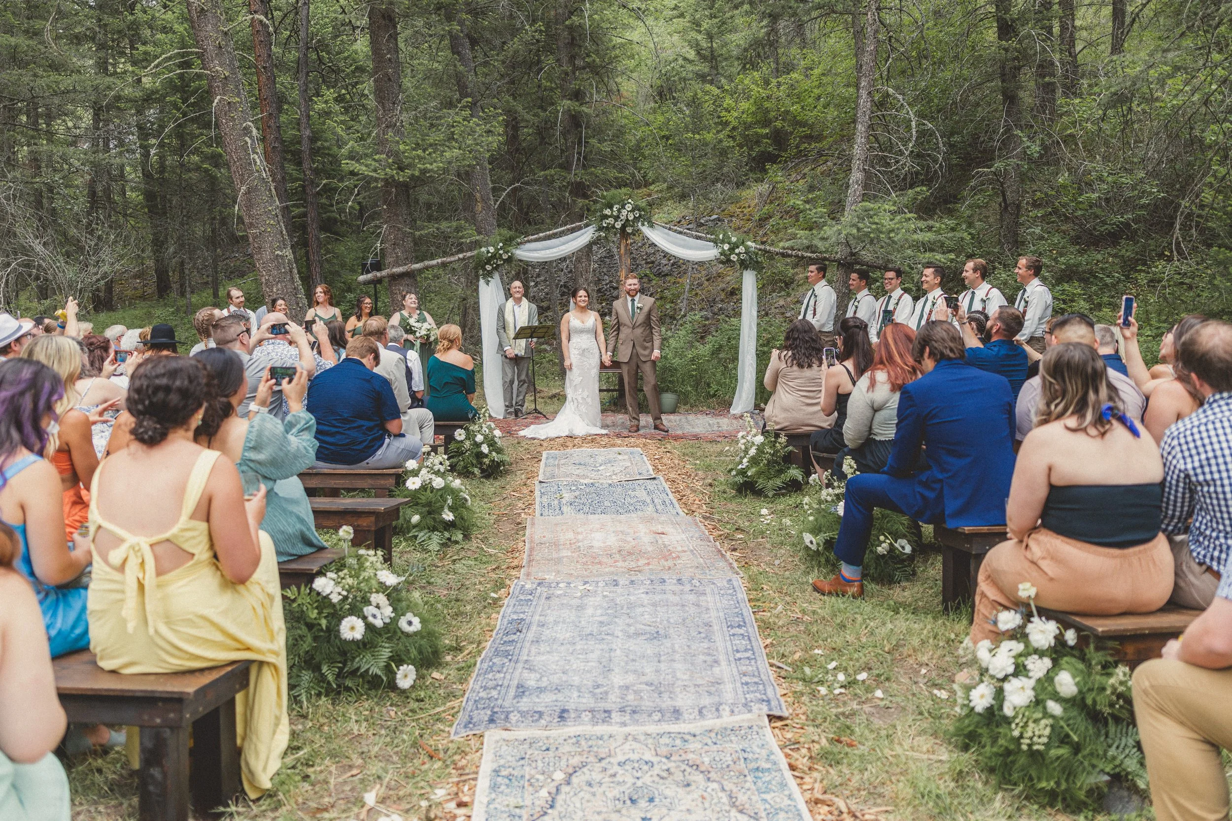 Outdoor wedding ceremony in a wooded area with a bride and groom standing under a decorated arch, surrounded by guests seated on benches, some taking photos, with a musical group on the right side.