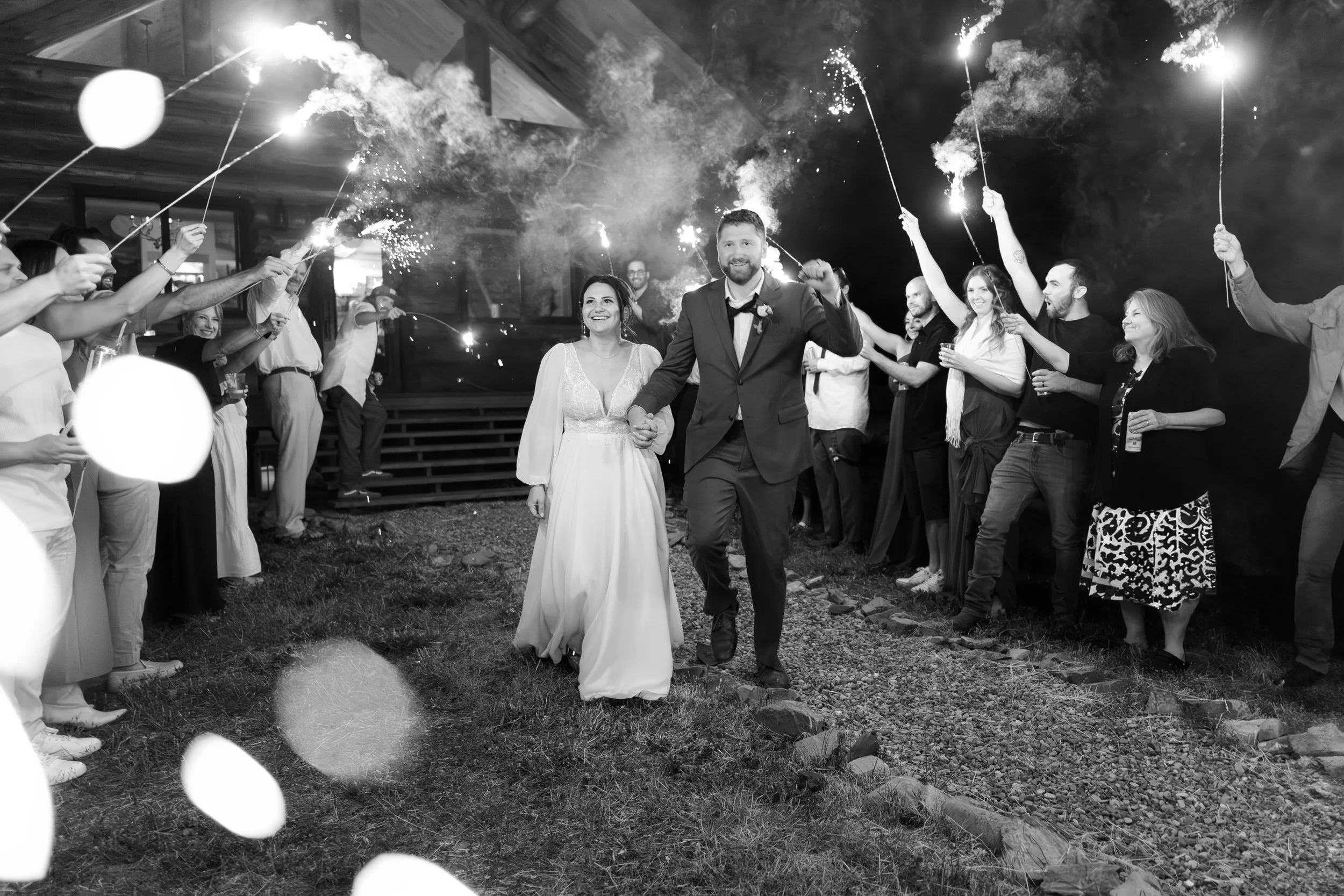 A wedding celebration with a bride and groom walking hand-in-hand through a line of guests holding sparklers at night.
