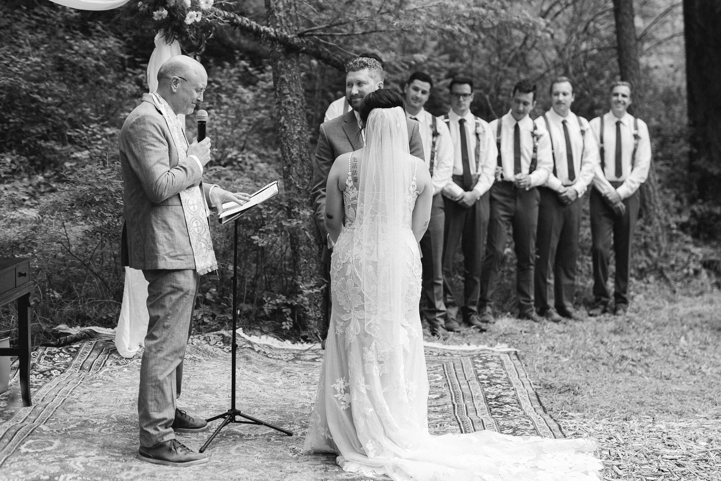 A black and white photo of a wedding ceremony outdoors. The bride and groom are exchanging vows in front of an officiant holding a microphone and reading from a book. The bride wears a long, lace wedding gown with a veil, and the groom is in a suit. 