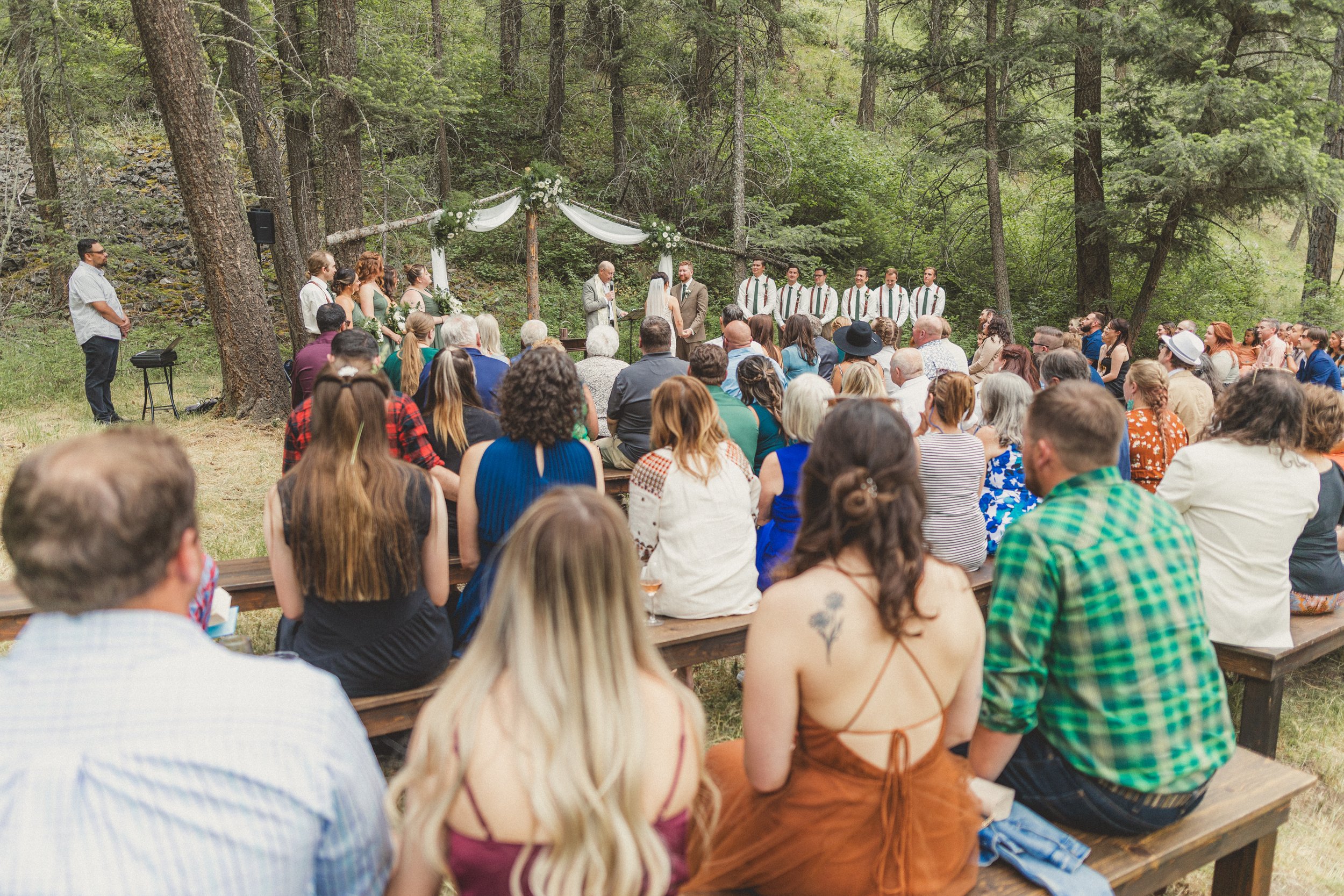 Outdoor wedding ceremony in a forest with guests seated on benches and a couple standing before an officiant under a decorated arch.
