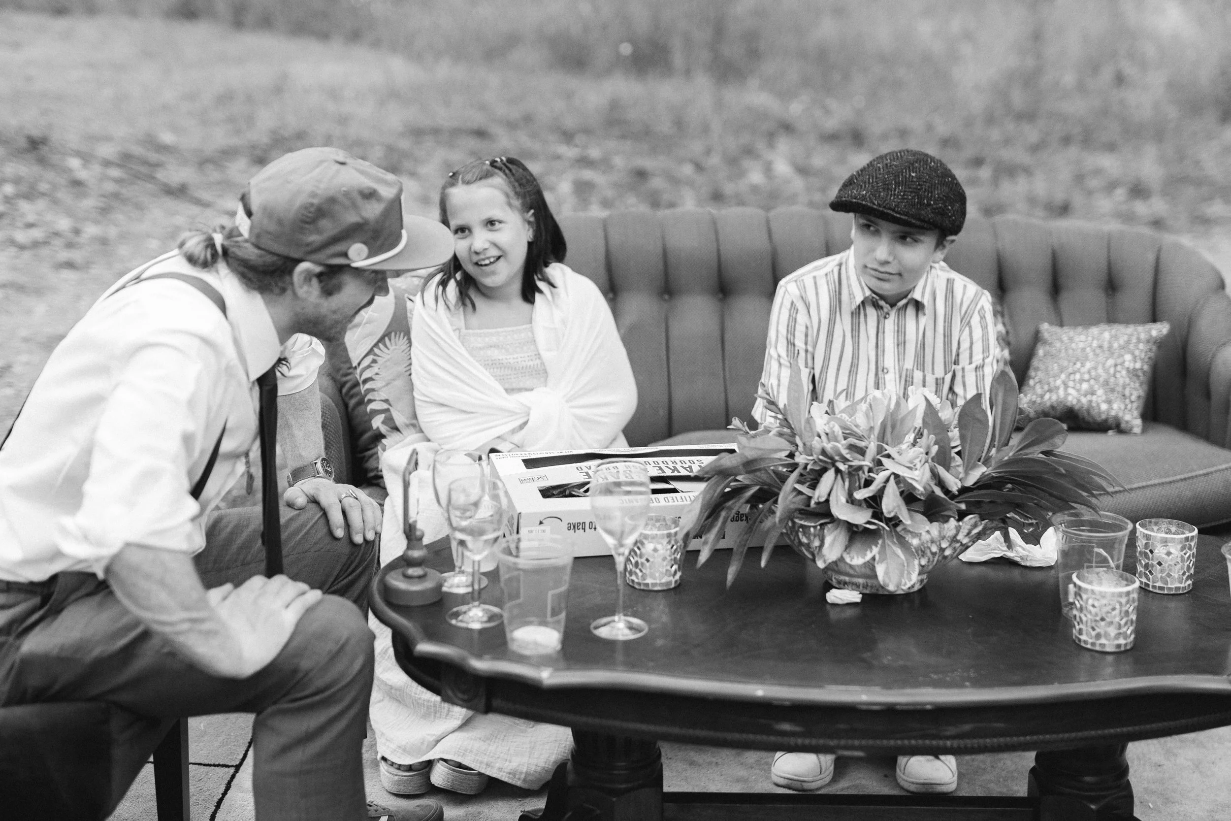 Three children sitting on a sofa outside at a table with drinks and a large floral arrangement, engaging with an adult who is leaning in to talk to them.