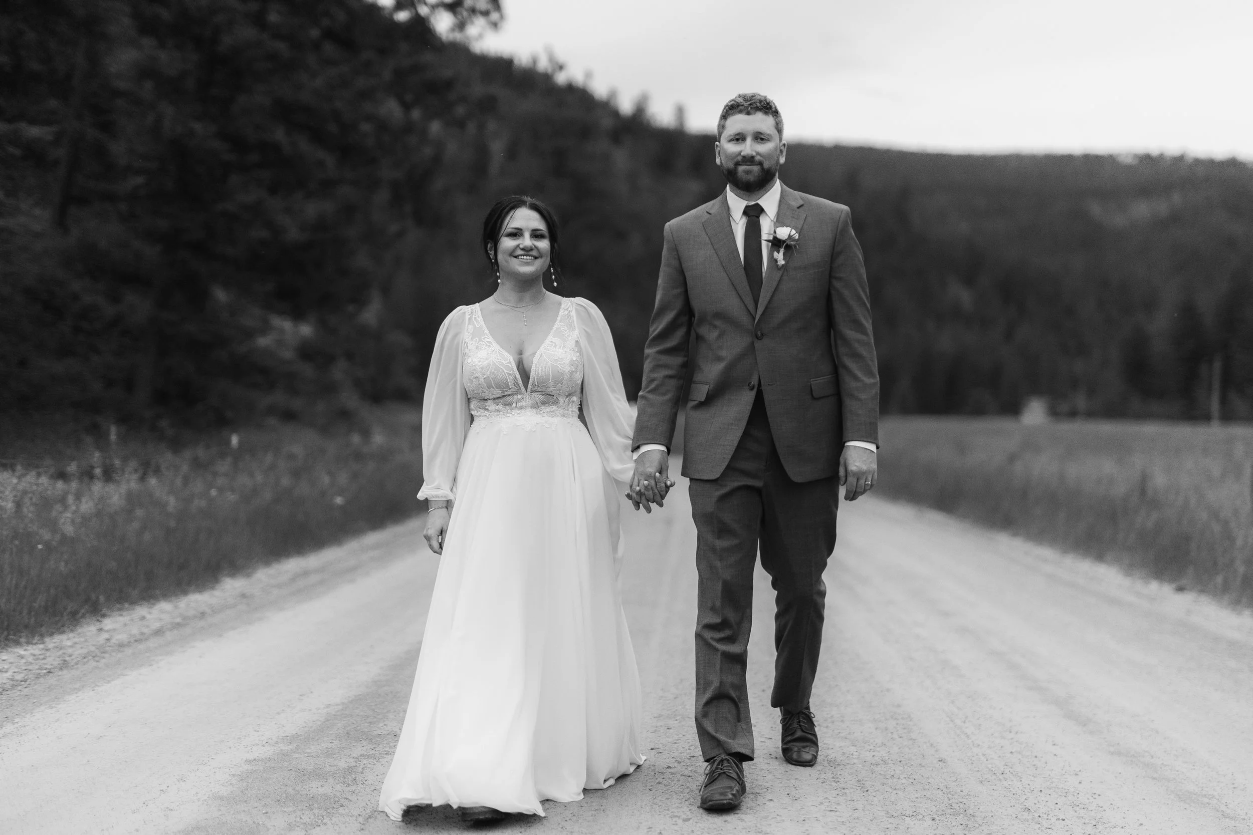 Black and white photo of a newly married couple walking hand in hand on a dirt road surrounded by grass and trees. The bride is wearing a long white wedding dress with lace details and the groom is wearing a tailored suit with a boutonniere.