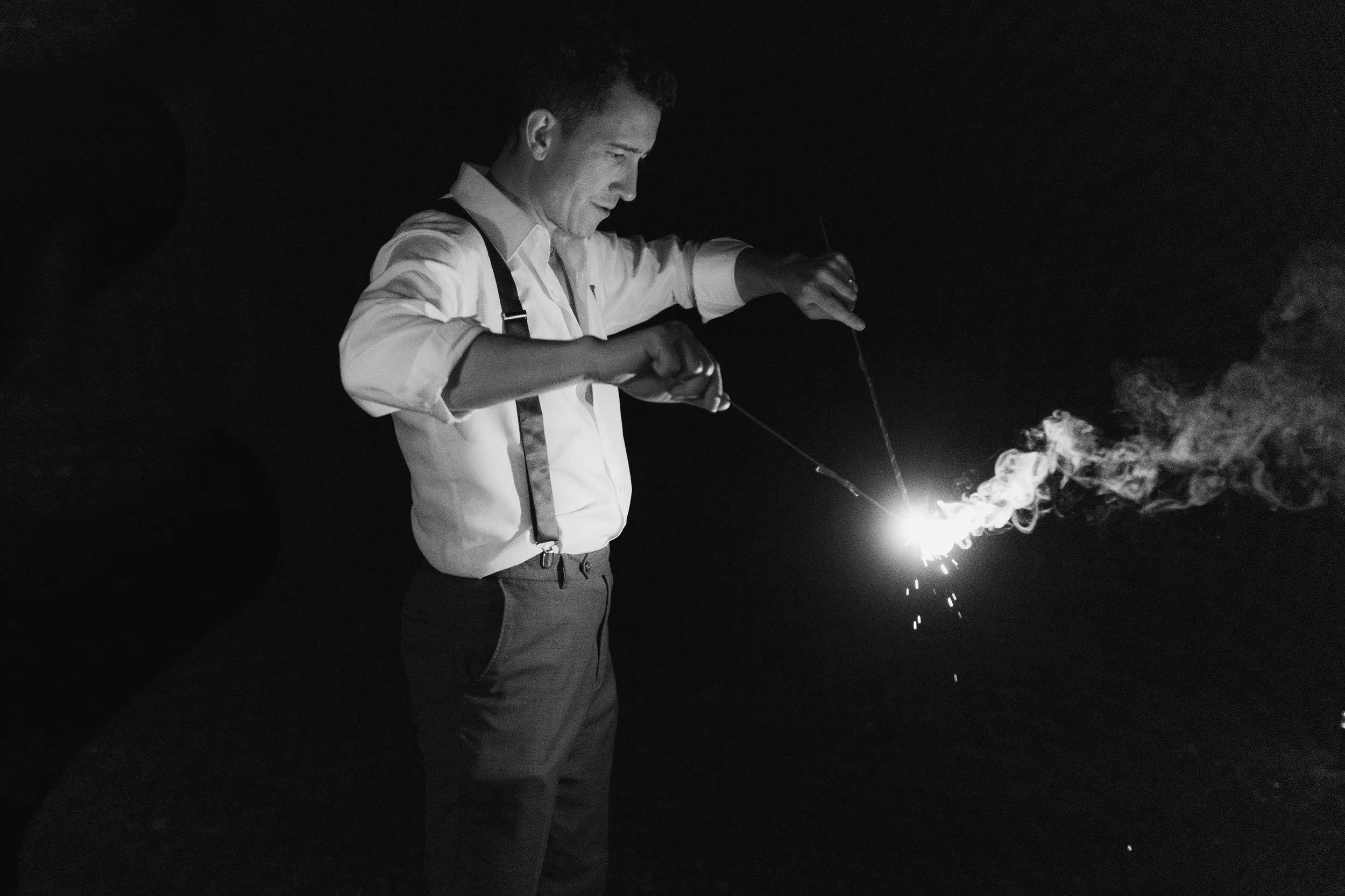 A man is holding a sparkler in each hand, with smoke and sparks visible, in a dark setting.