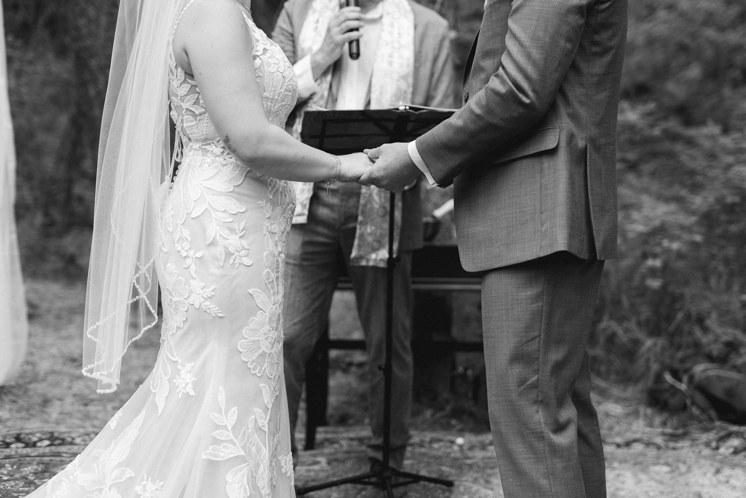 A black and white photo of a couple holding hands during a wedding ceremony, standing outdoors with an officiant officiating, with trees in the background.