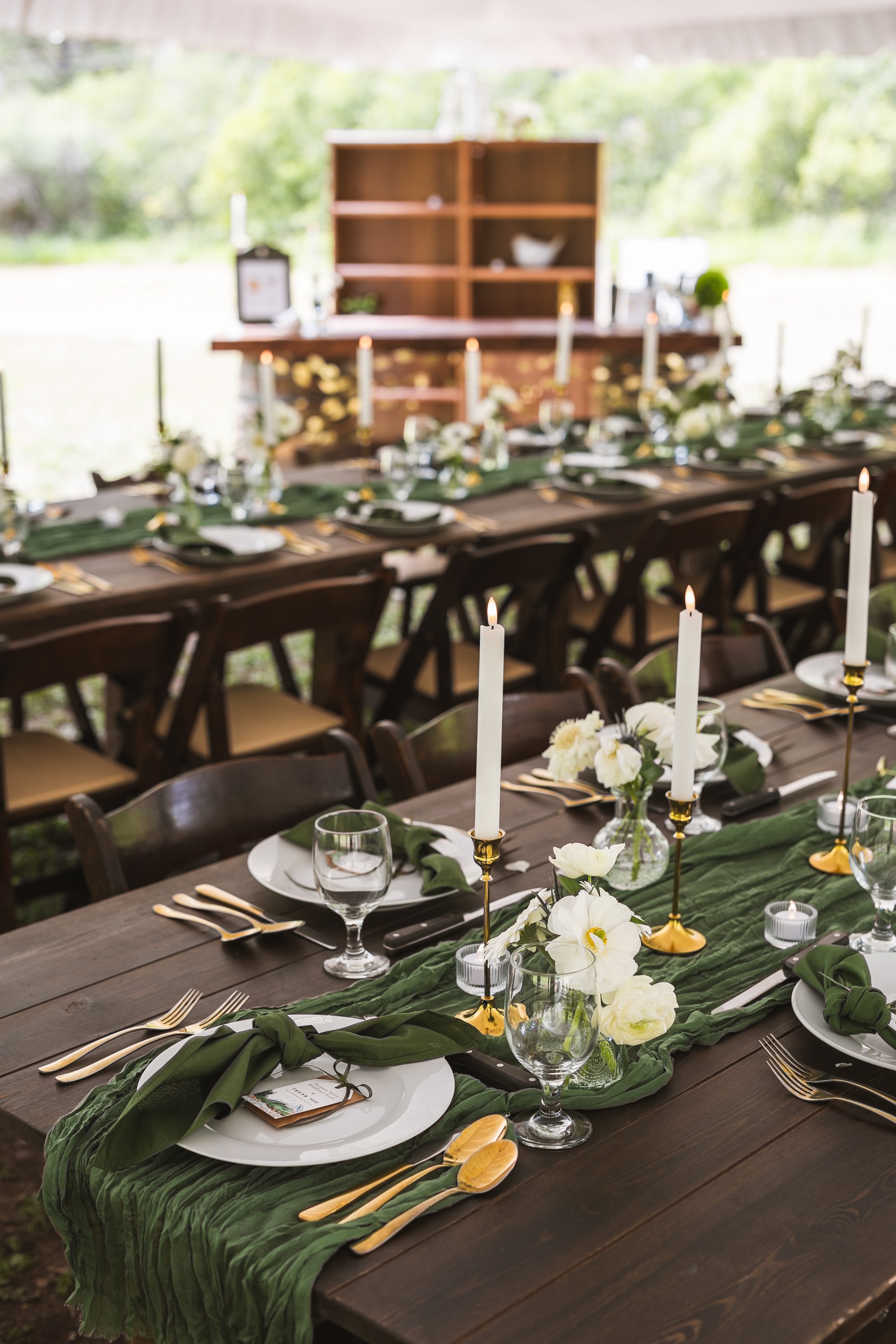 Decorated outdoor dining table with white flowers, tall white candles, gold cutlery, and green table runner.
