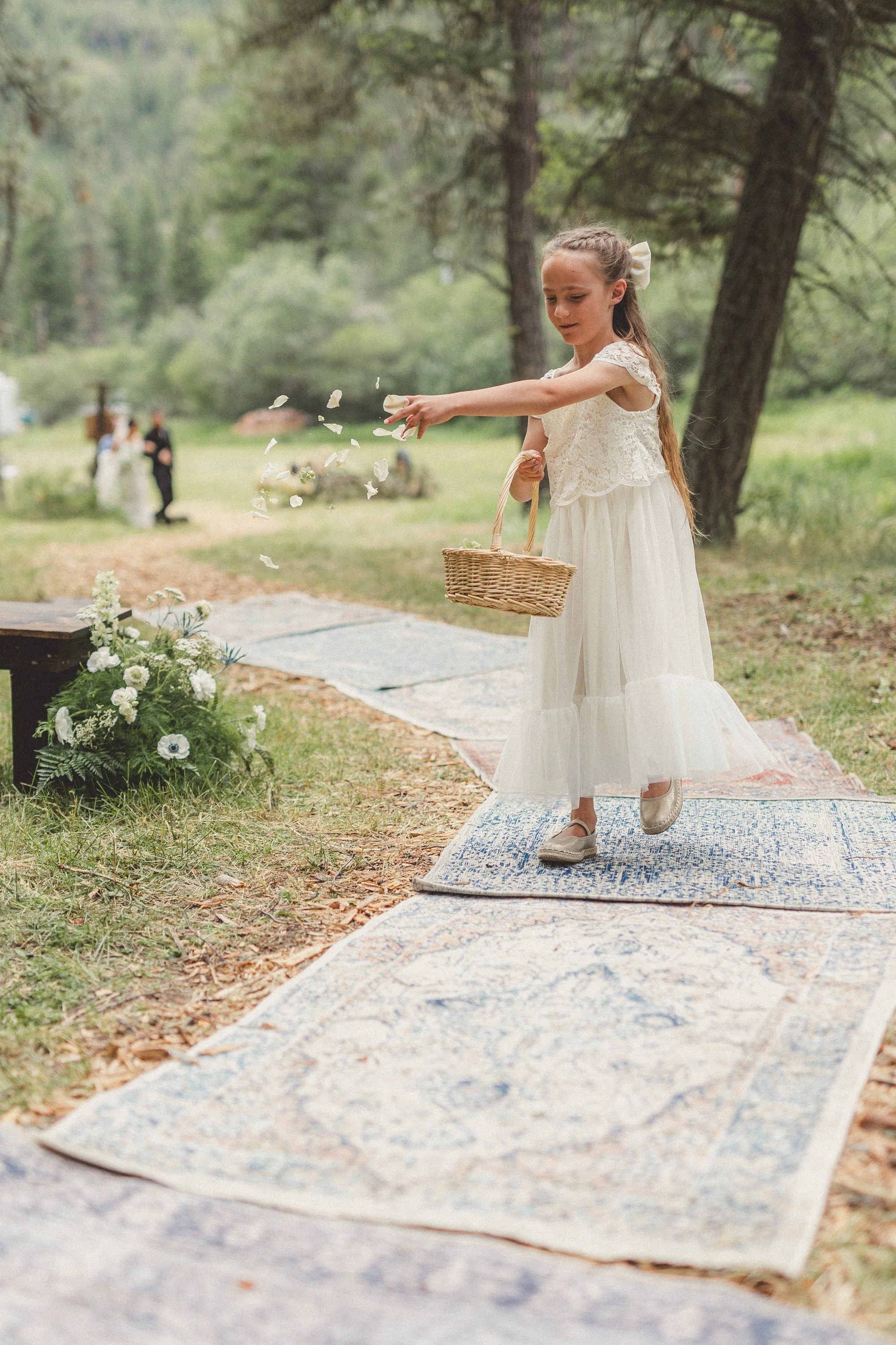 A girl in a white dress and silver shoes throwing flower petals on a patterned outdoor rug during a wedding ceremony in a wooded area.