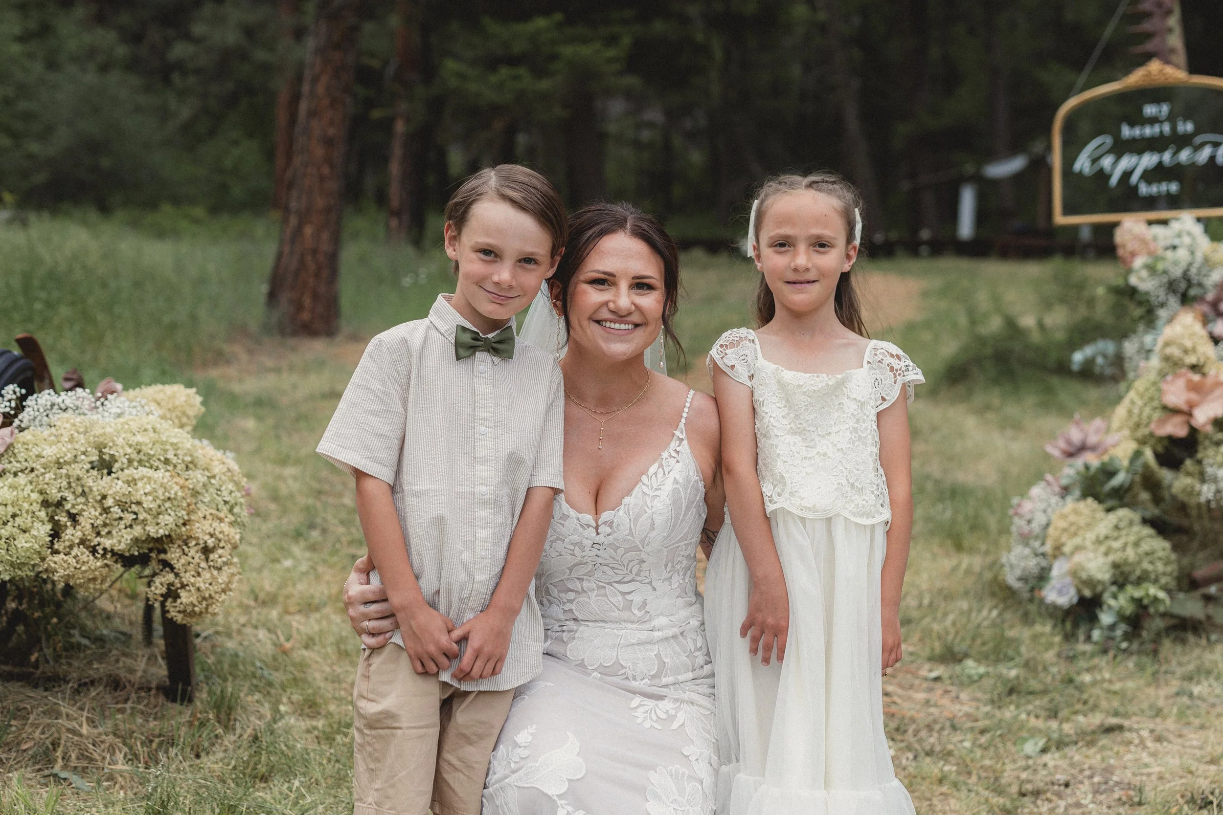 A woman in a wedding dress posing with two children, a boy and a girl, at an outdoor wedding ceremony. They are standing on grass, with trees and floral decorations in the background.