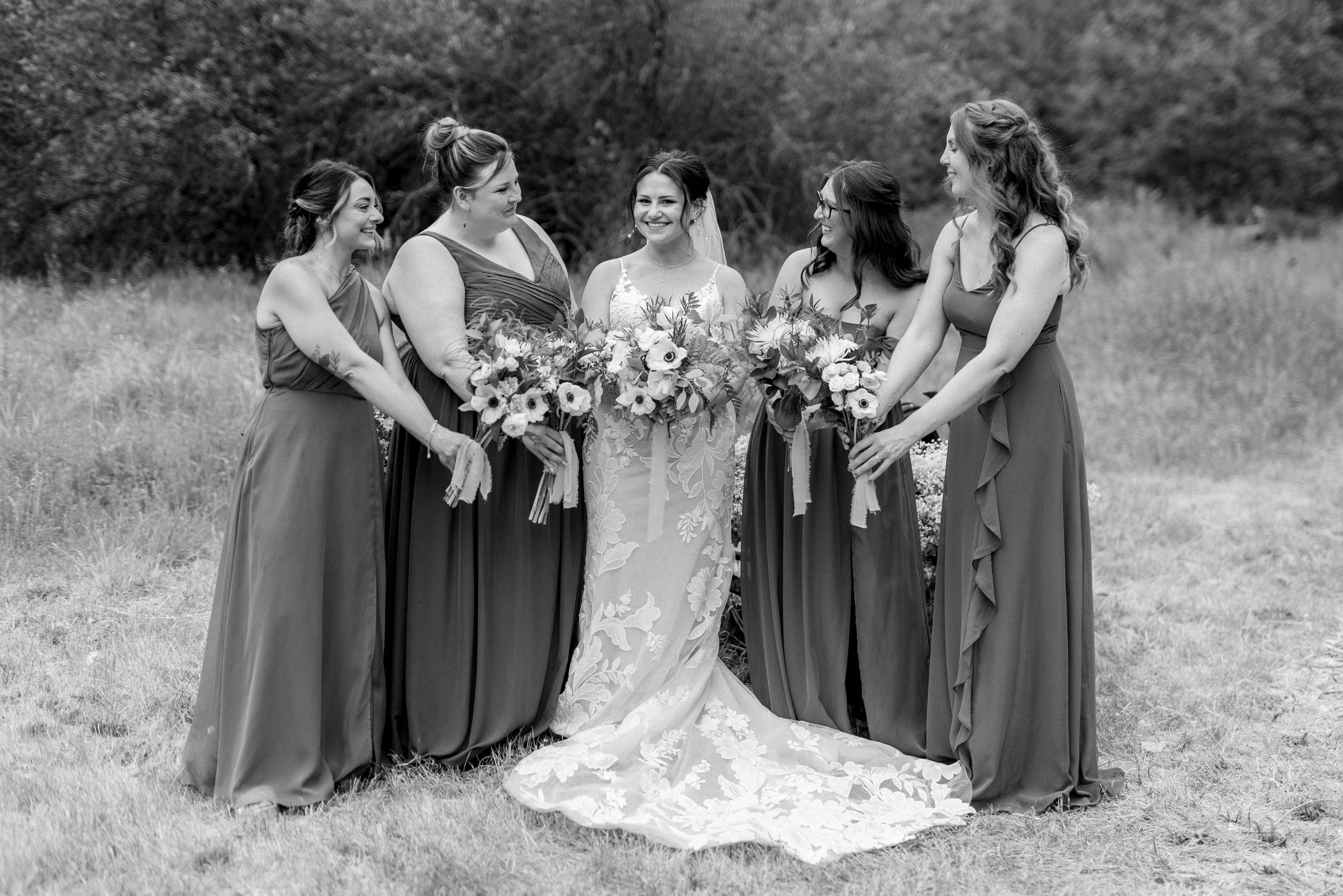 A bride and five bridesmaids standing outdoors on grass, smiling and holding bouquets of flowers.