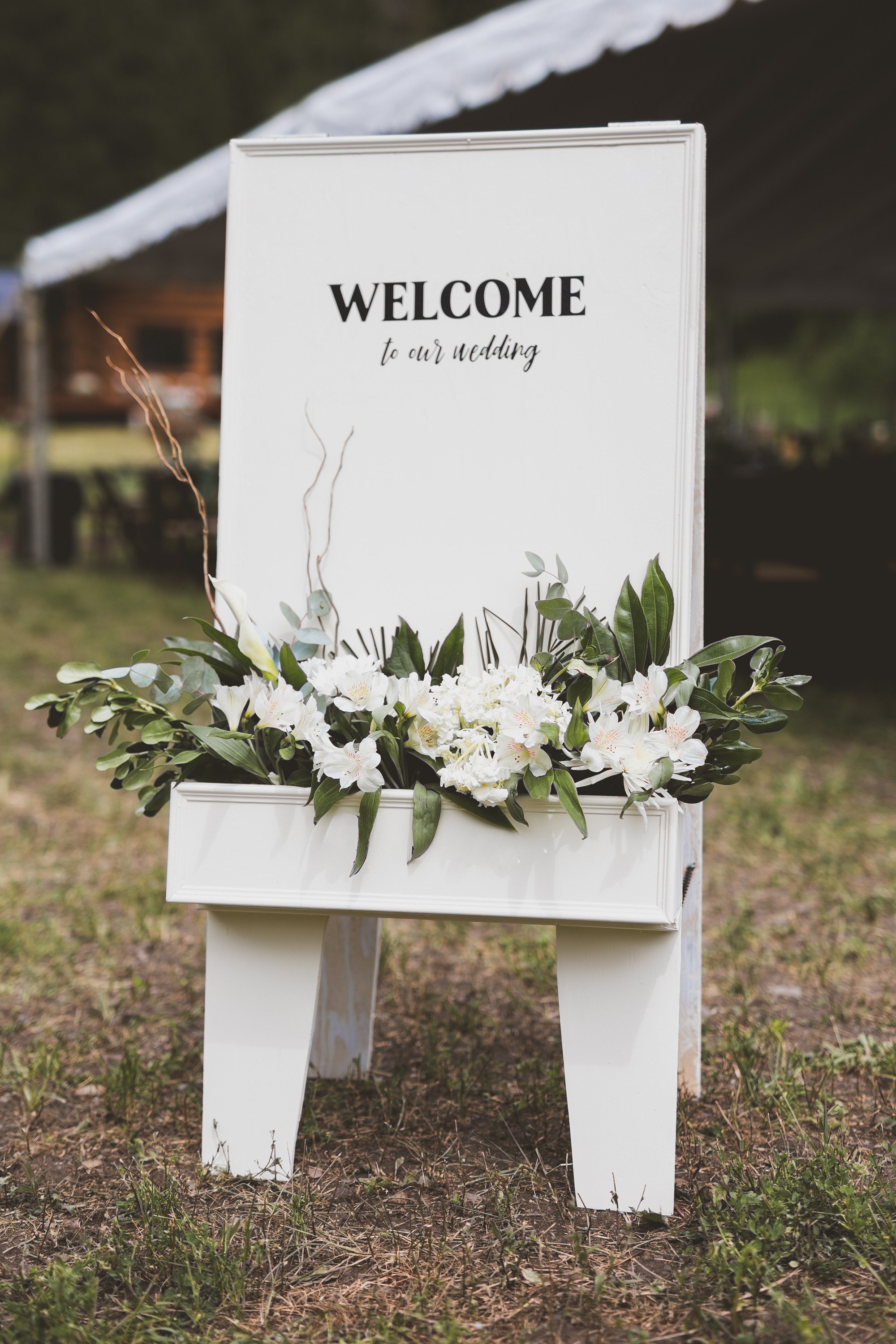 White wedding welcome sign with floral decoration at the bottom, placed outdoors on grass.
