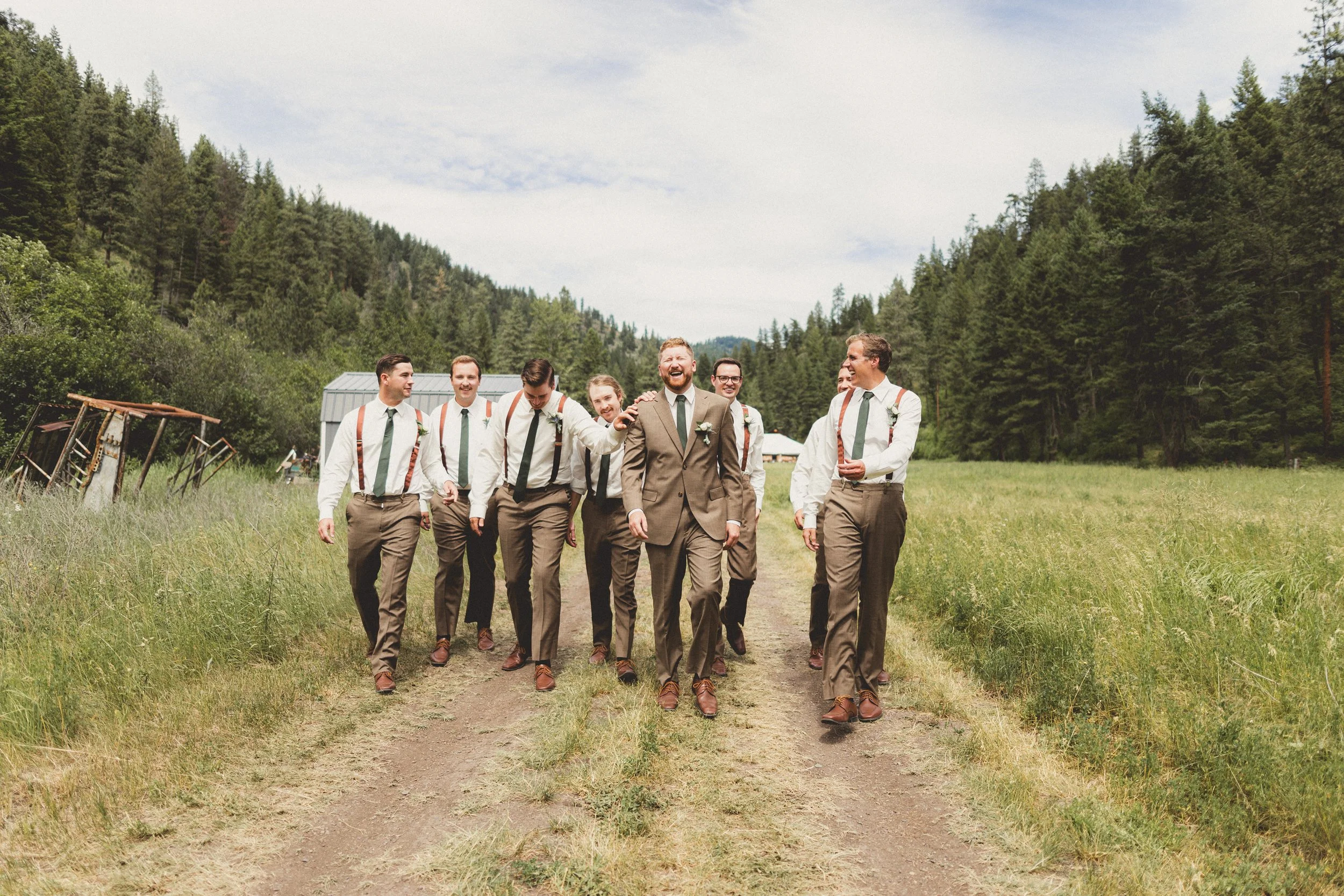 Group of men in formal attire walking on a dirt path in a grassy field with trees in the background, enjoying conversation.