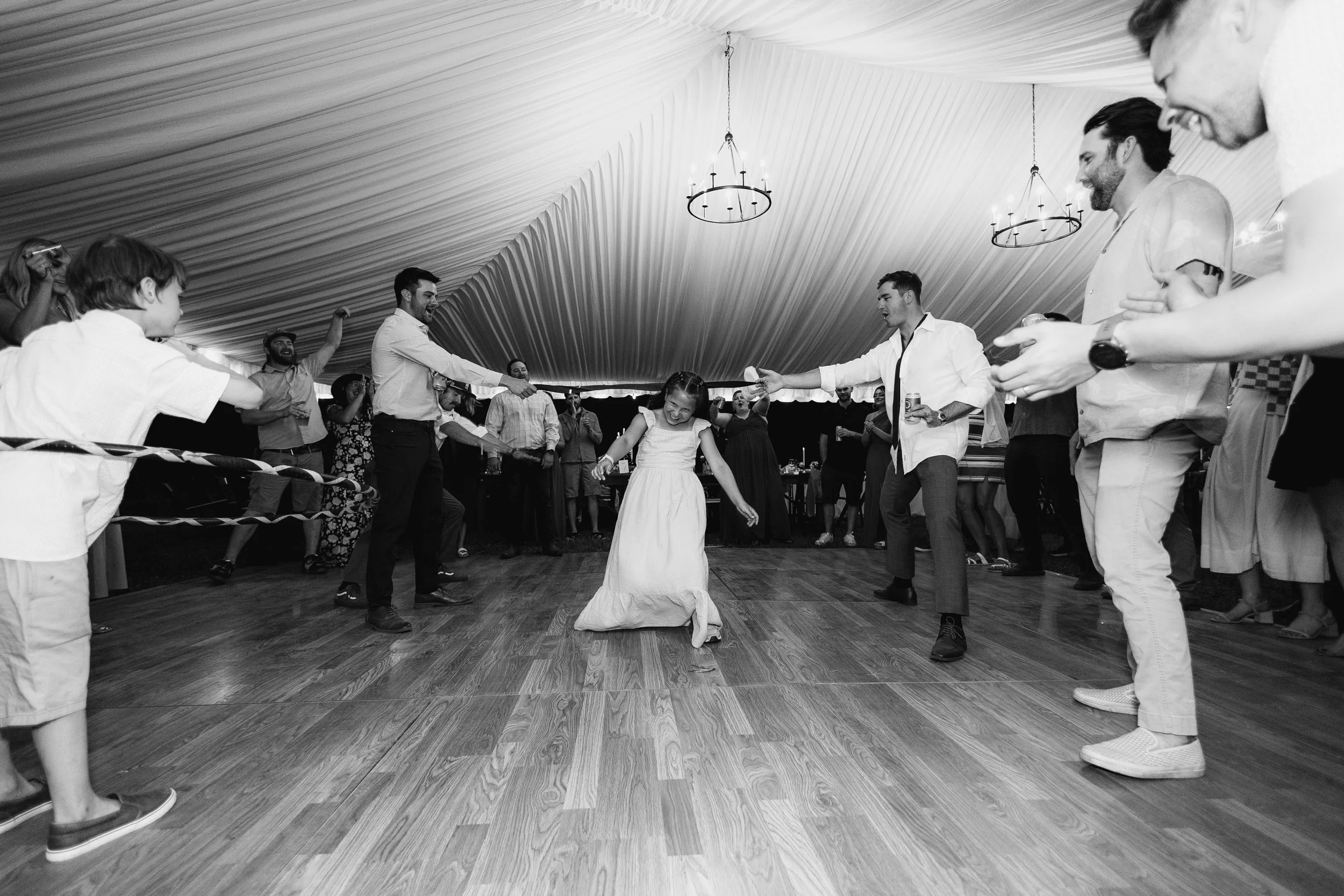 A young girl in a white dress dancing in the center of a circle of people holding hands at a celebration or wedding reception in a decorated tent.