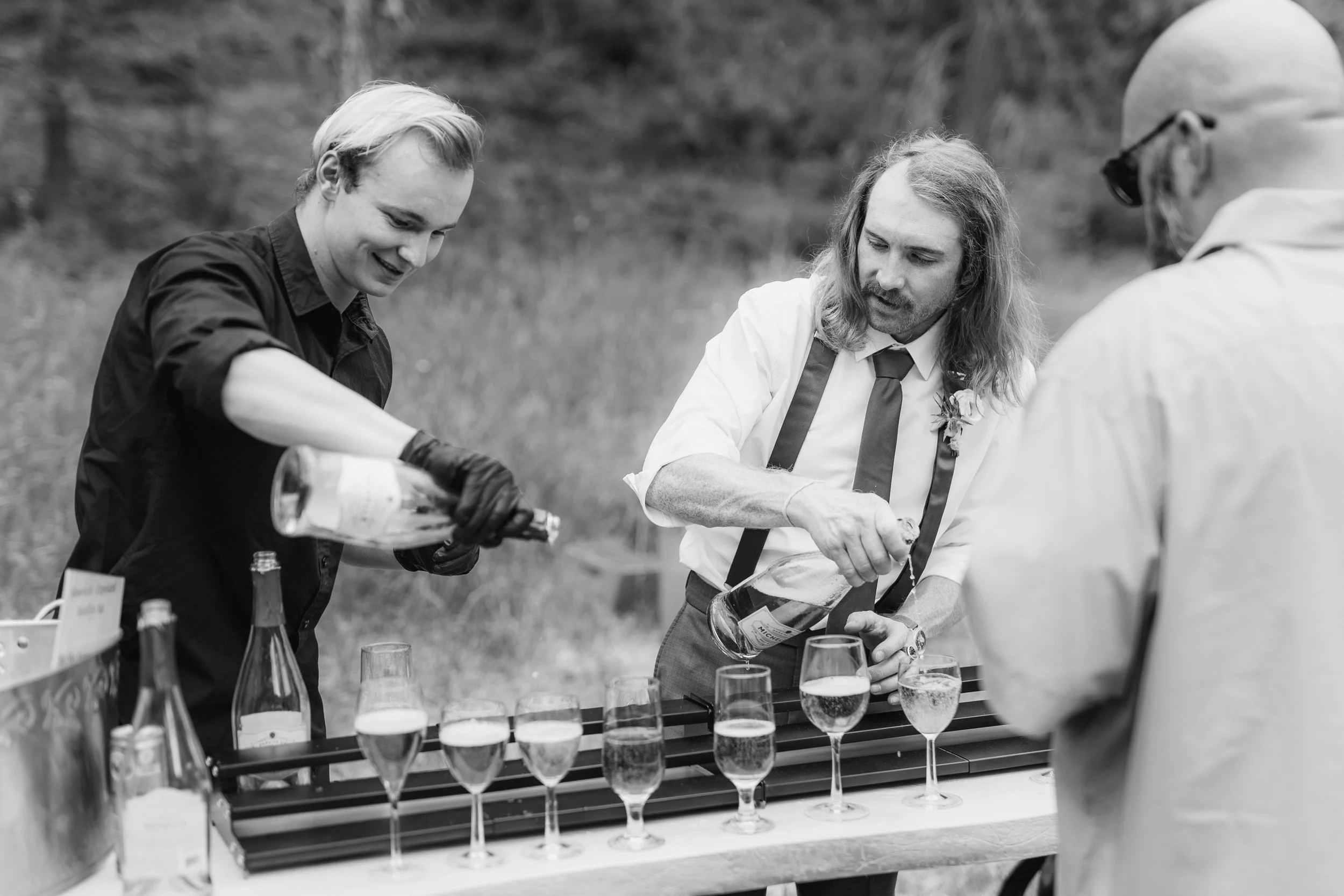 Two women and a man pouring drinks at an outdoor event, with glasses lined up on a table, black and white photo.