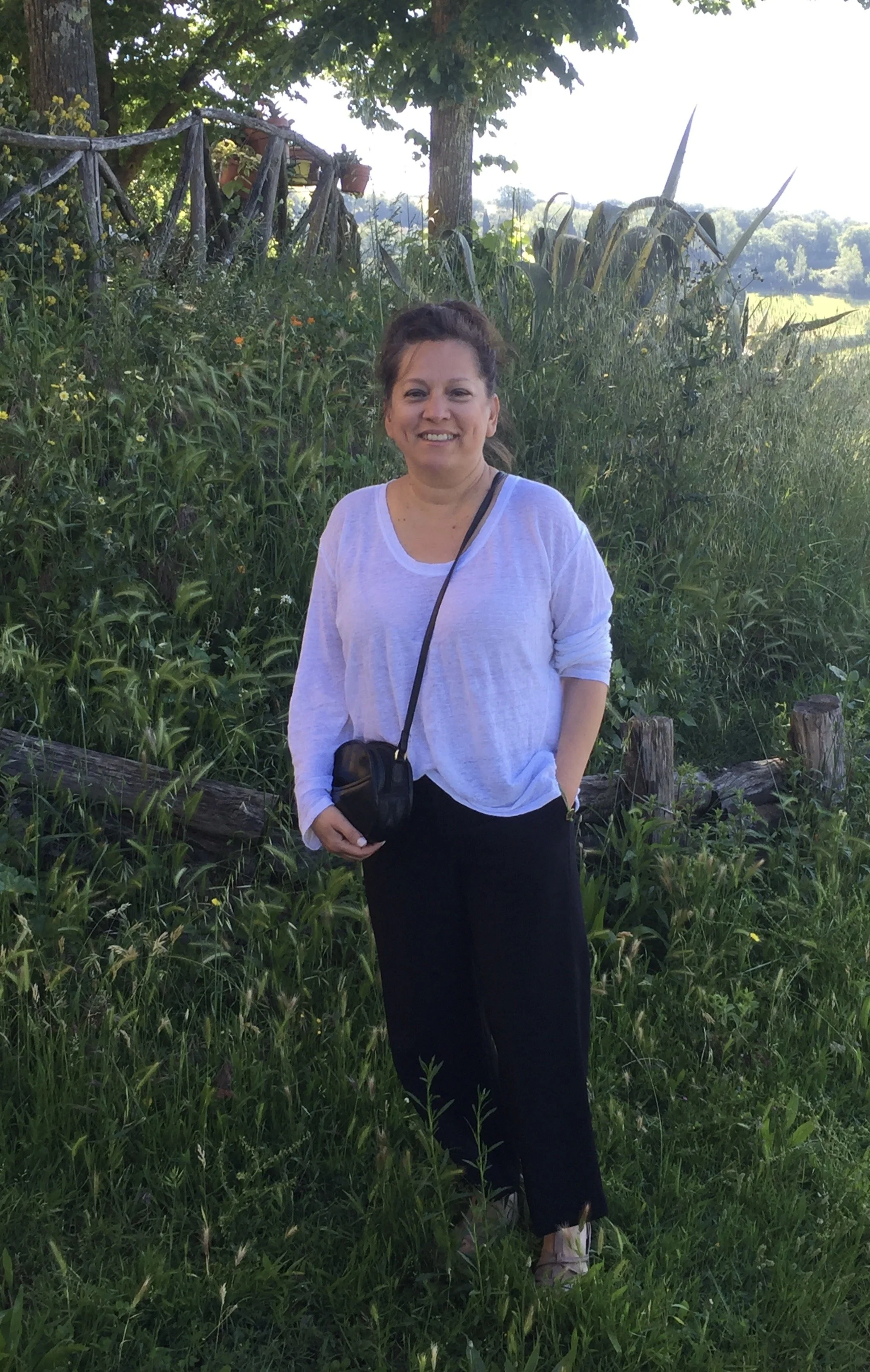 A woman standing outdoors in front of greenery, wearing a white long-sleeve top, black pants, and holding a black bag, smiling at the camera.