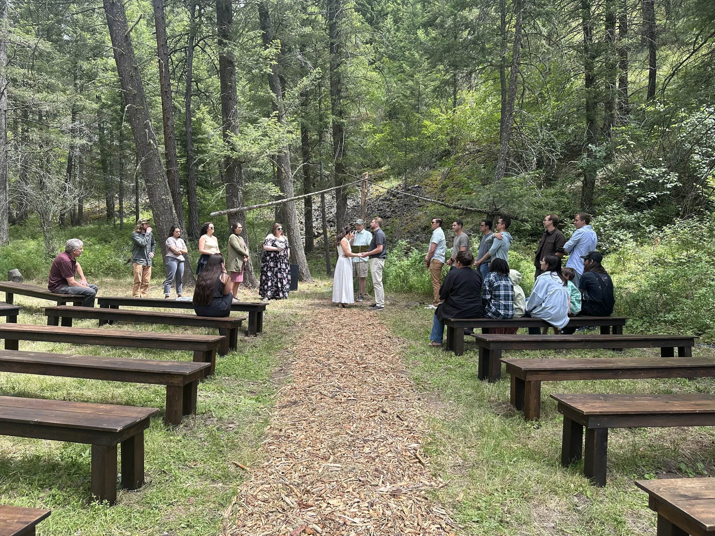 A group of people gathered in a forest for a wedding ceremony, with some seated on wooden benches and others standing, facing two people exchanging vows.