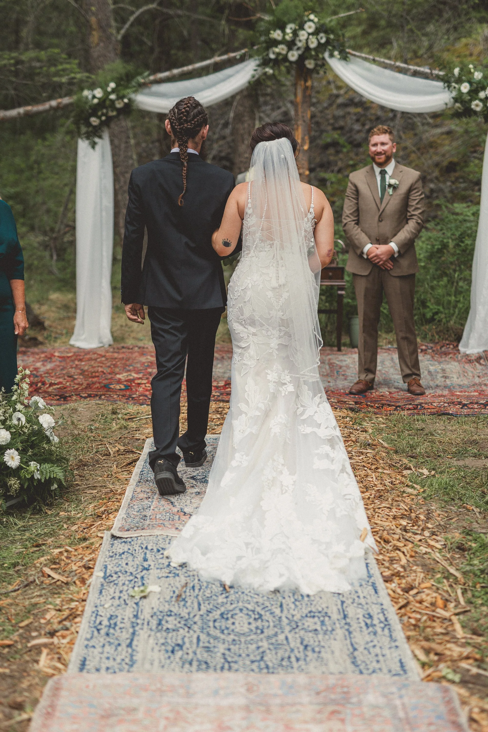 A same-sex couple getting married outdoors during their wedding ceremony, walking down the aisle towards an officiant, with a decorated arch and forest background.