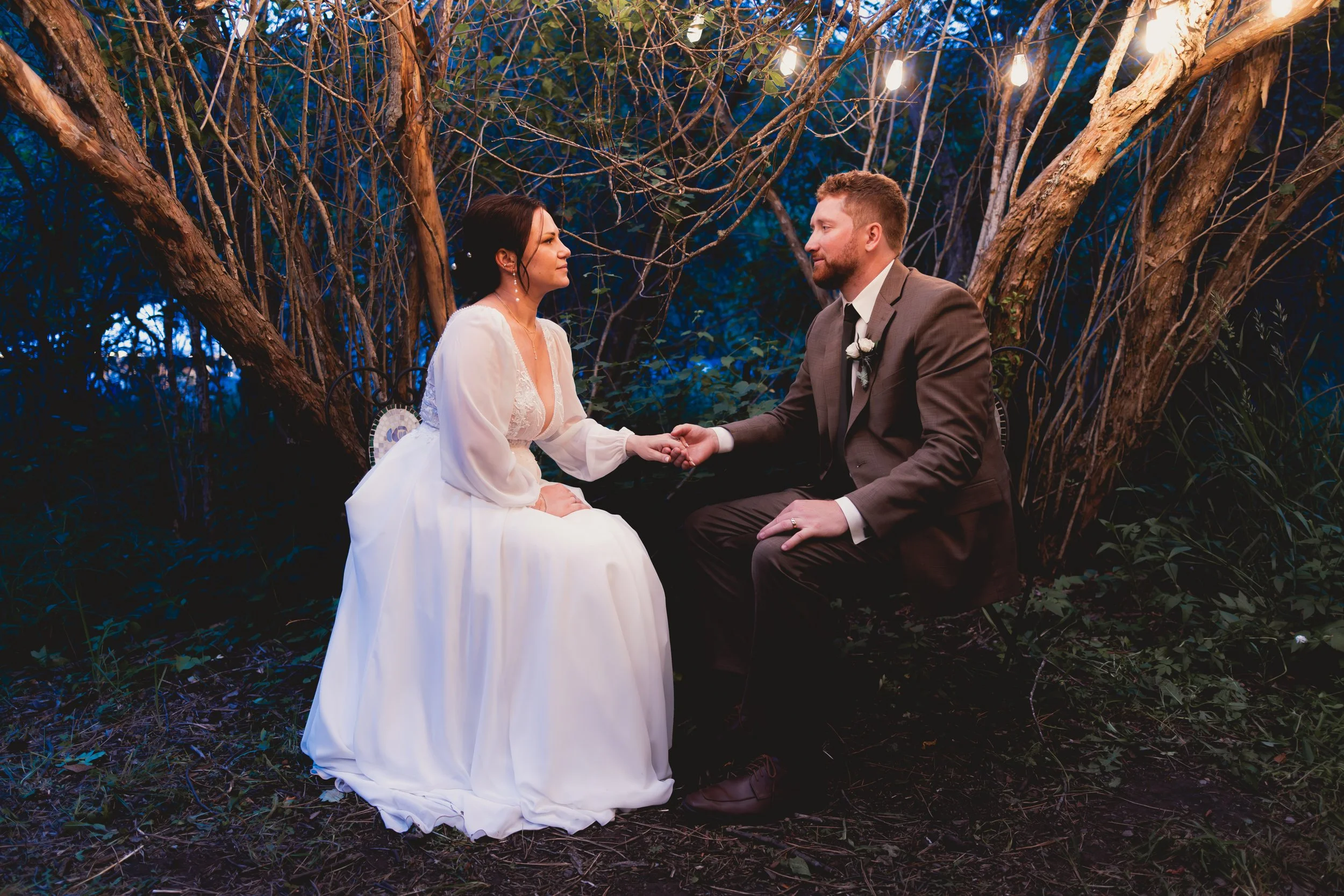 A bride and groom holding hands and gazing at each other in an outdoor setting at night, surrounded by trees with hanging lights.