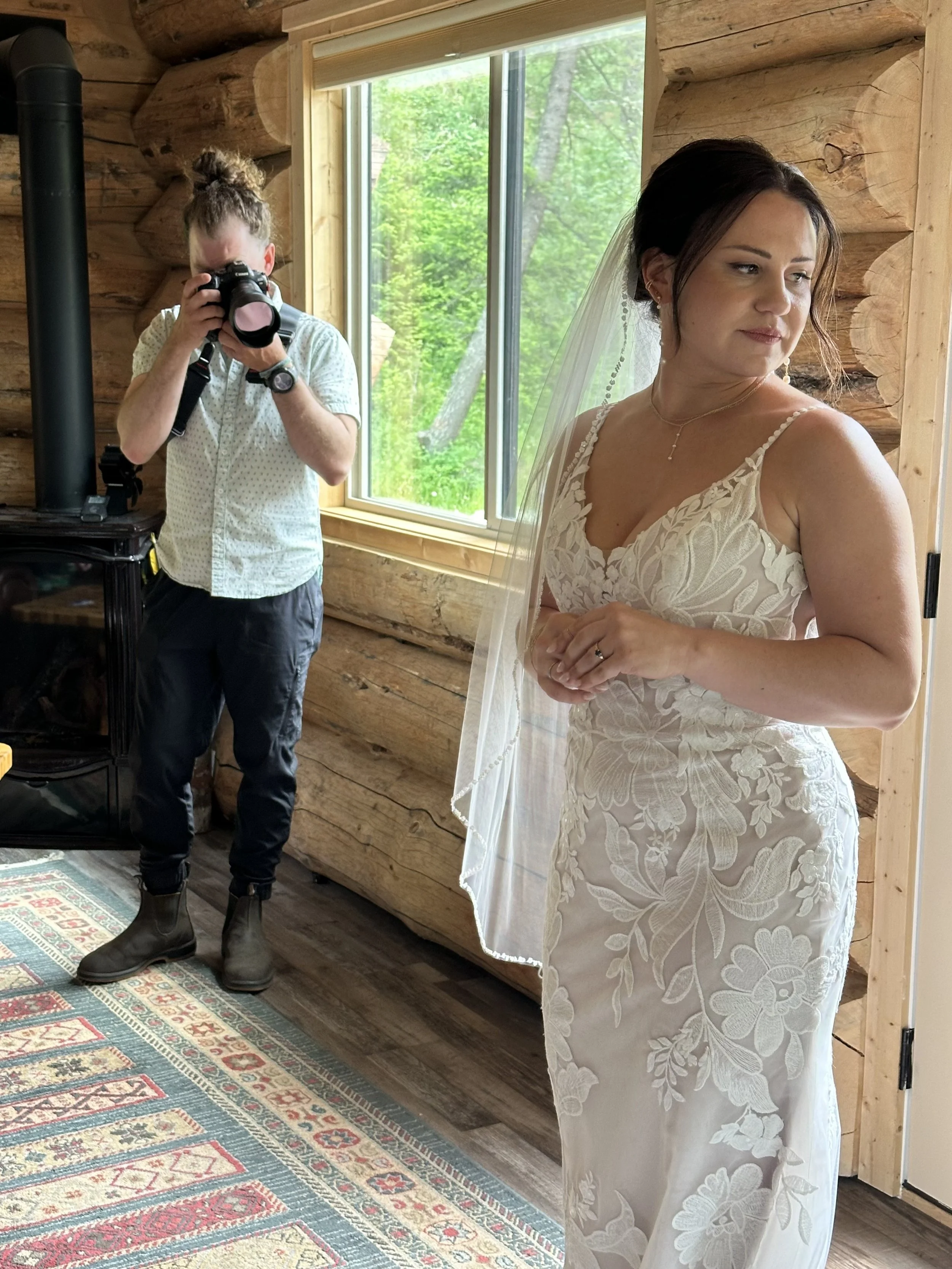 A bride in a floral lace wedding dress and veil stands indoors, looking to the side, with a photographer taking her picture in a rustic wooden room with a window showing greenery outside.