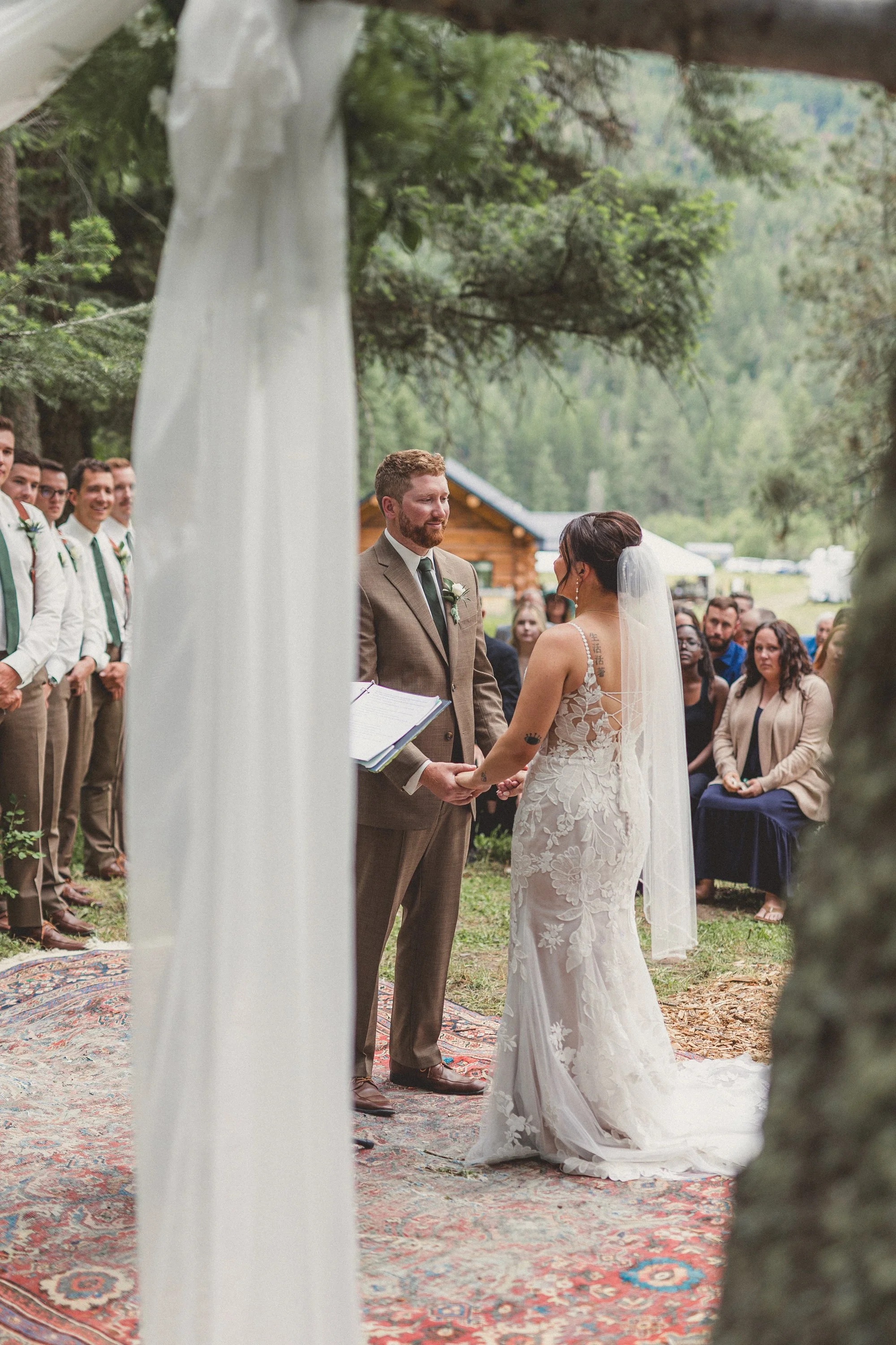 A bride and groom exchanging vows outdoors, surrounded by wedding guests, trees, and a rustic log building, during a wedding ceremony.