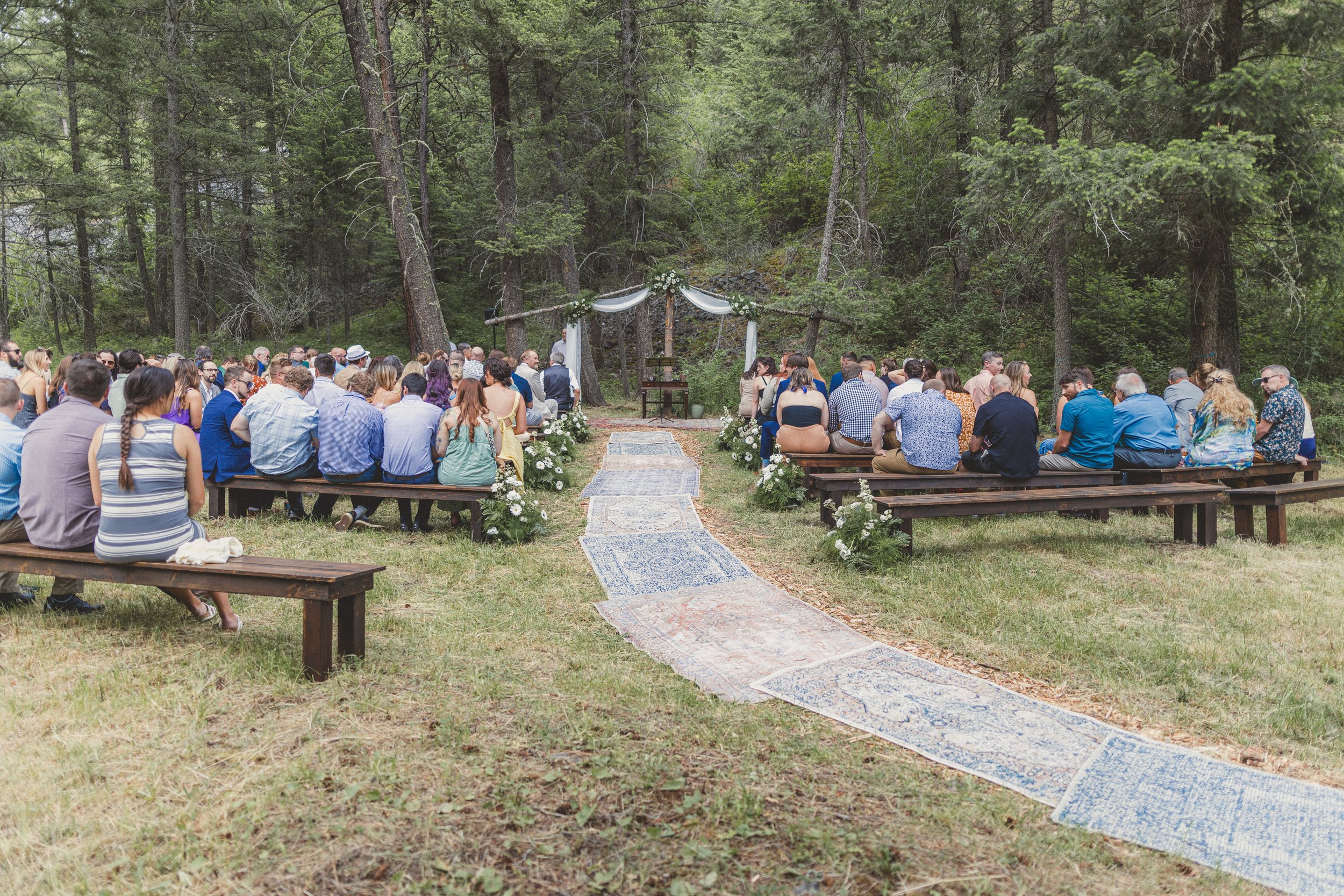 Outdoor wedding ceremony with guests seated on benches in a forest setting, decorated with flowers and drapes.