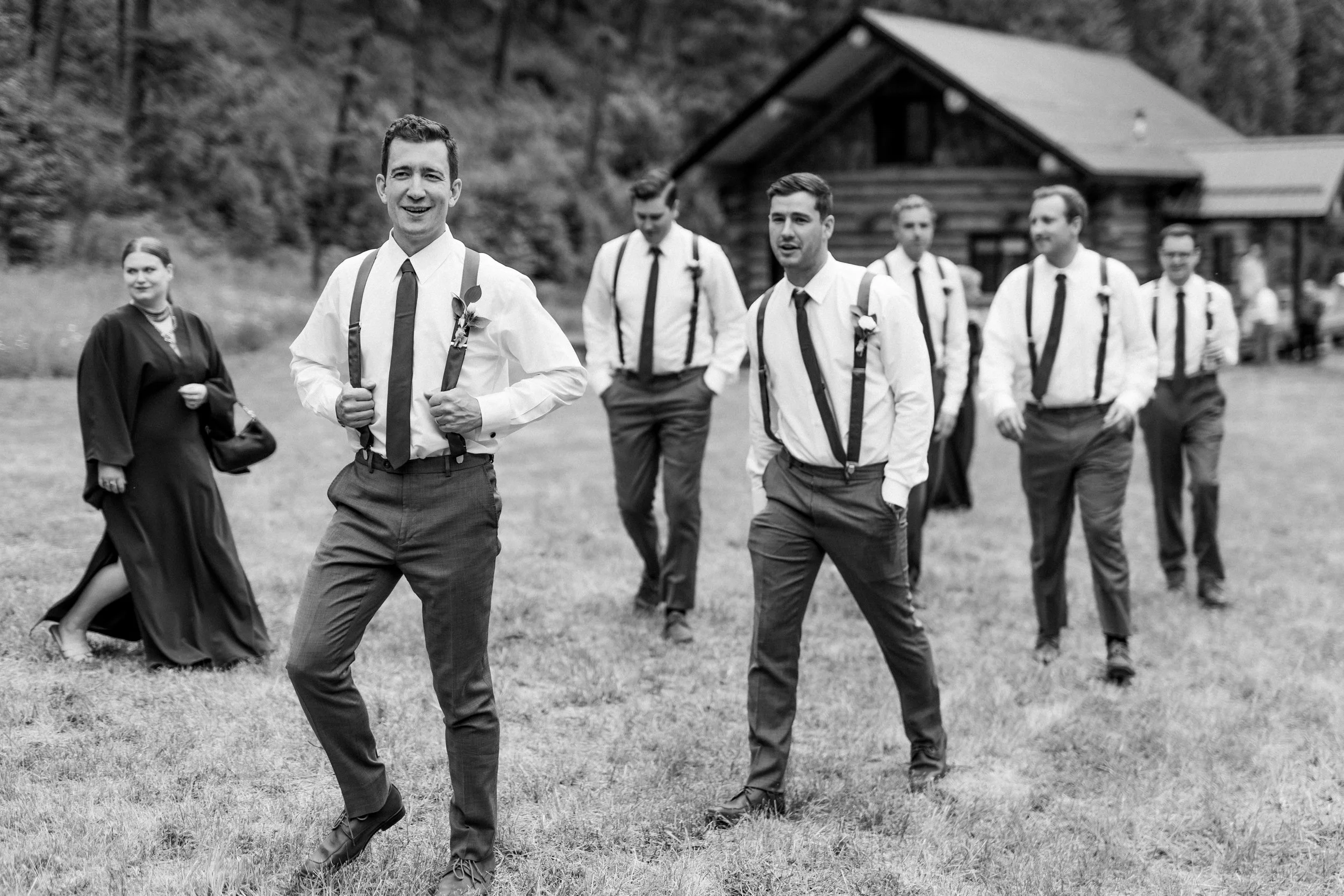 A group of men and a woman in wedding attire, walking on a grassy area outdoors near a wooden cabin. The men are dressed in white shirts with suspenders and ties, and the woman is wearing a black dress with a sash, holding a purse.