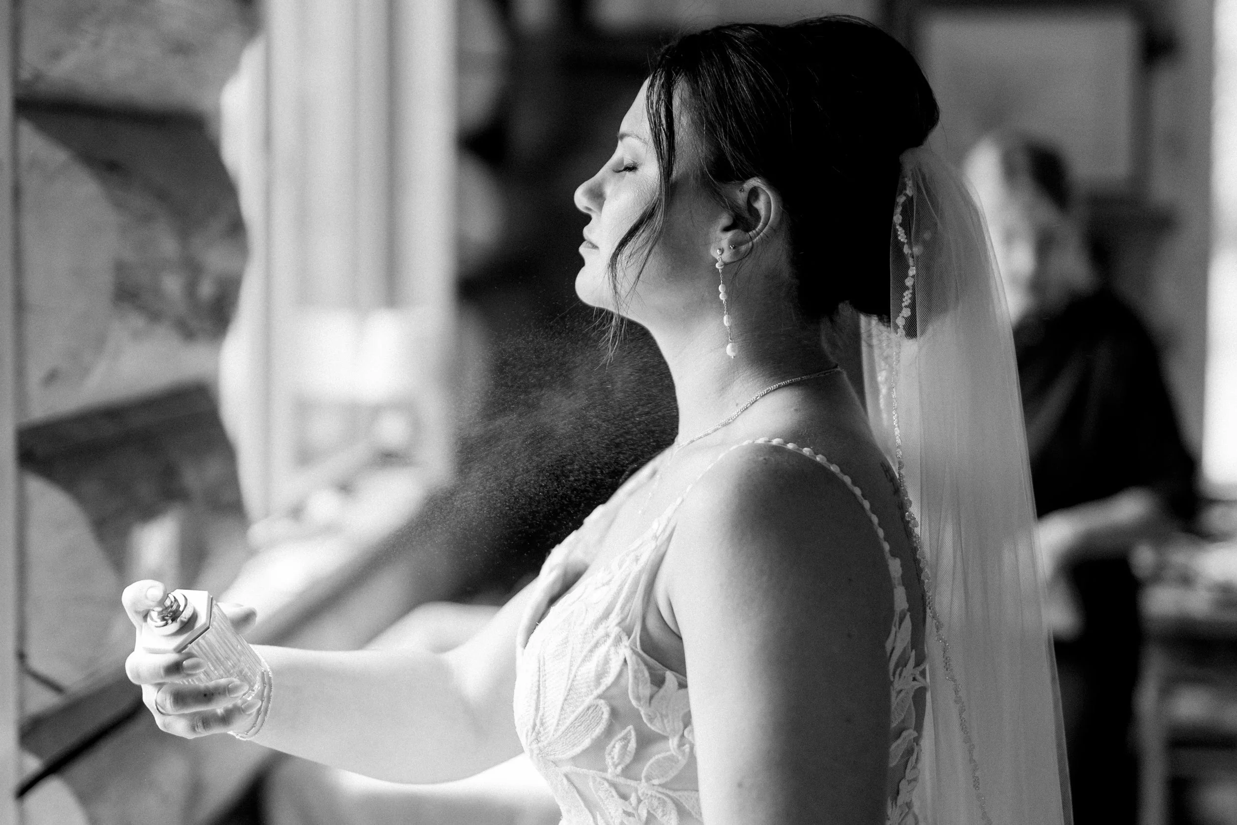 Bride with veil and earrings sprays perfume onto her neck in a black-and-white photo.